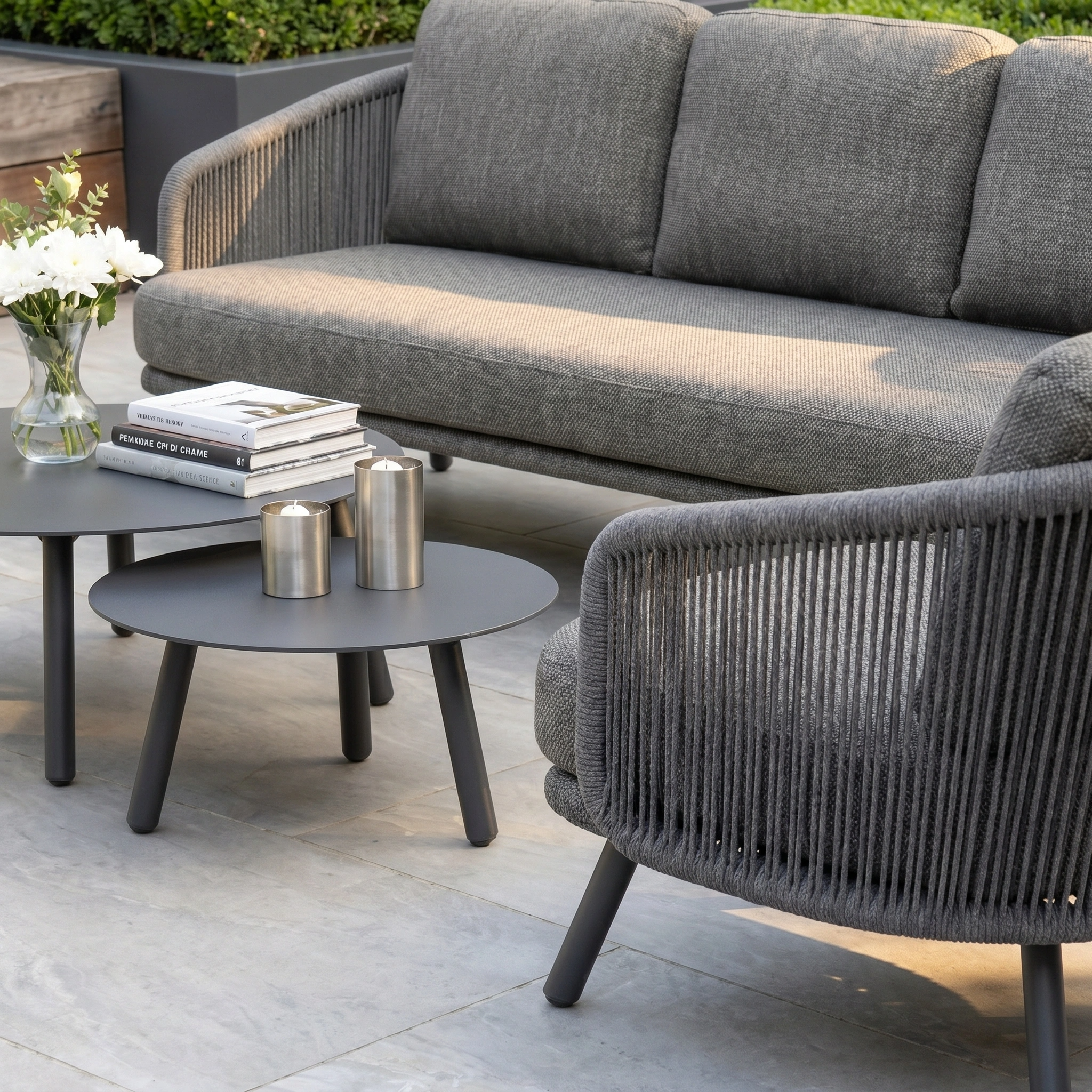 The Dekota 3 Seat Rope Sofa Set with Coffee Tables and Footstool in Charcoal features stylish outdoor accents, including an olefin rope weave, enhanced with white flowers and candles.