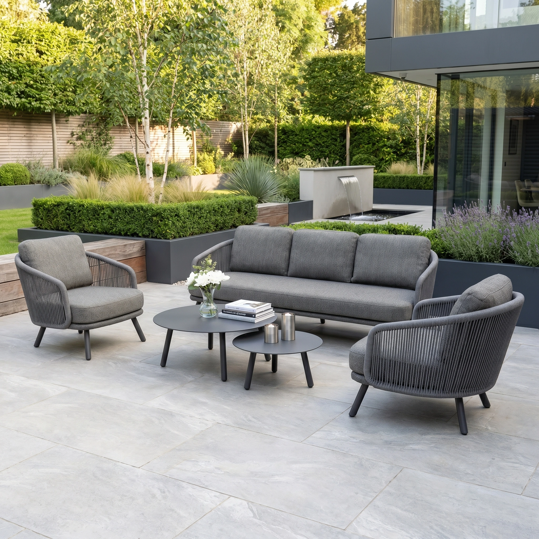 Modern patio with a Dekota 3 Seat Rope Sofa Set in charcoal, including armchairs, two coffee tables, and a footstool, surrounded by lush greenery.