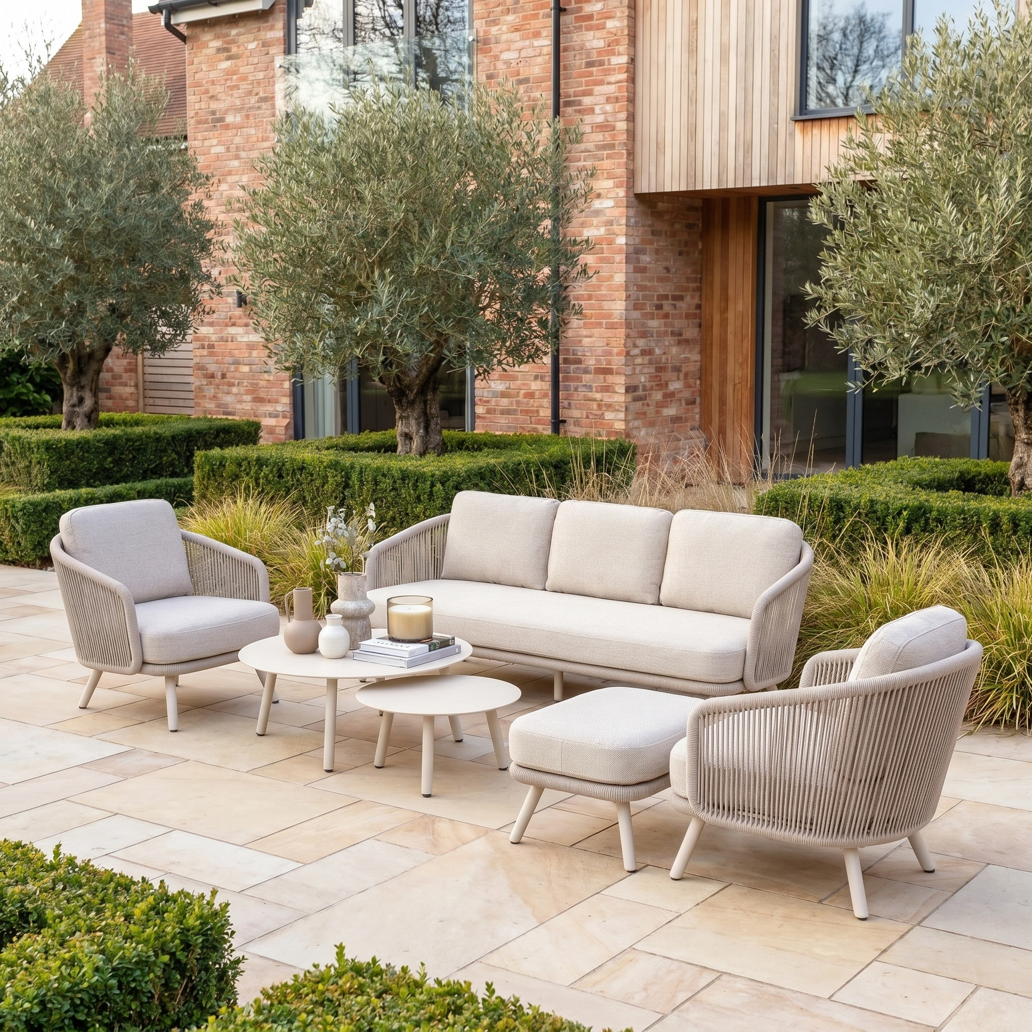 The Dekota 3 Seat Rope Sofa Set with Coffee Tables and Footstool in Fawn, upholstered in durable solution-dyed olefin fabric, offers lasting outdoor style on a patio accented by manicured hedges and olive trees.
