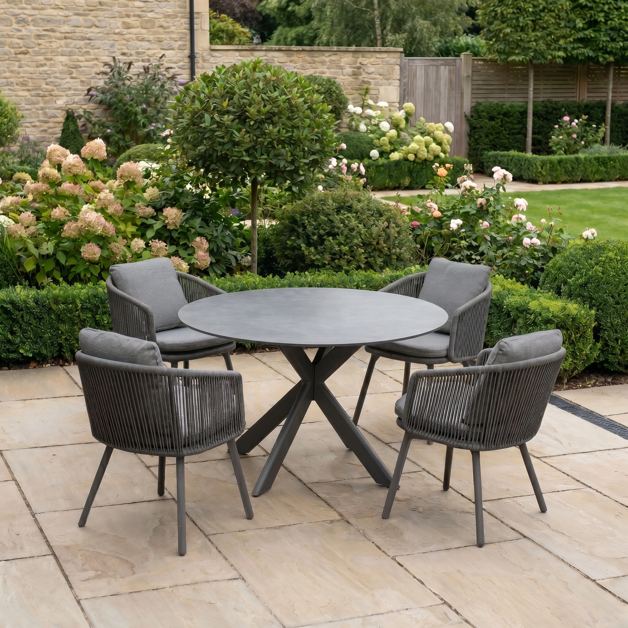 The Dekota 4 Seat Round Rope Dining Set with Albatross Table in Charcoal is ideal for stylish outdoor dining, set on a stone terrace and surrounded by a landscaped garden.