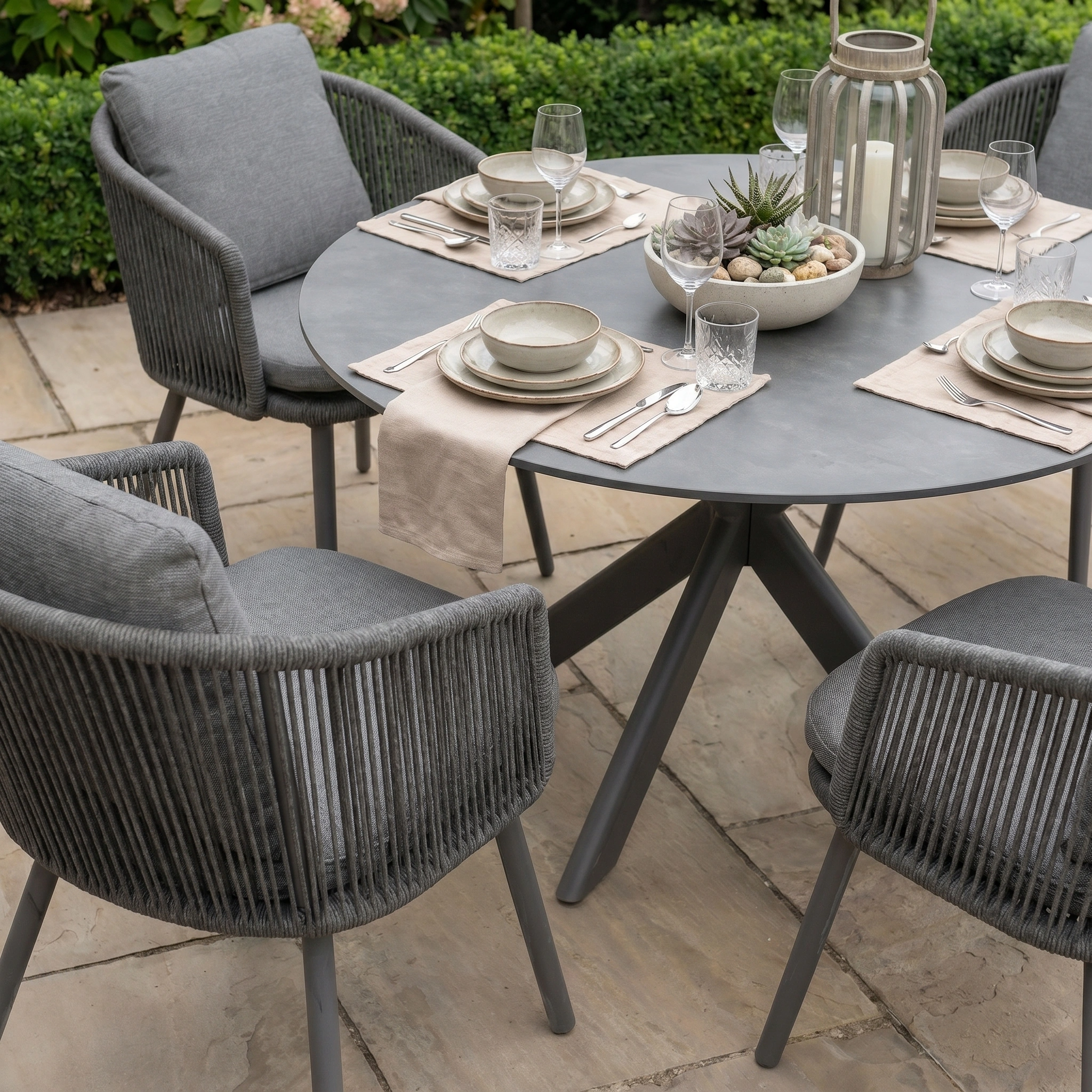 A grey rope chair is placed at the Albatross Table from the Dekota 4 Seat Round Rope Dining Set with Albatross Table in Charcoal, set with plates, glasses, and a succulent—ideal for stylish outdoor dining.