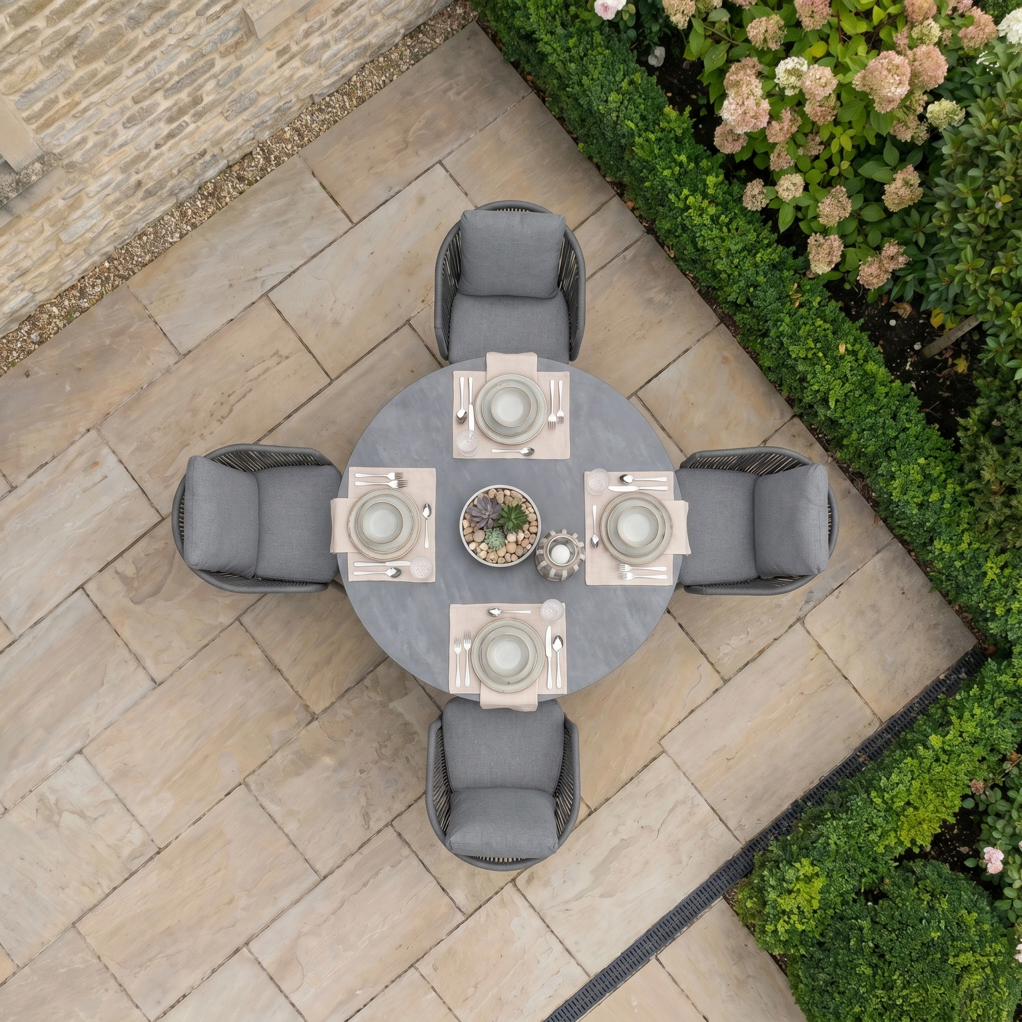 A Dekota 4 Seat Round Rope Dining Set with Albatross Table in Charcoal, viewed from above, is set among stone tiles and greenery—ideal patio furniture for outdoor gatherings.