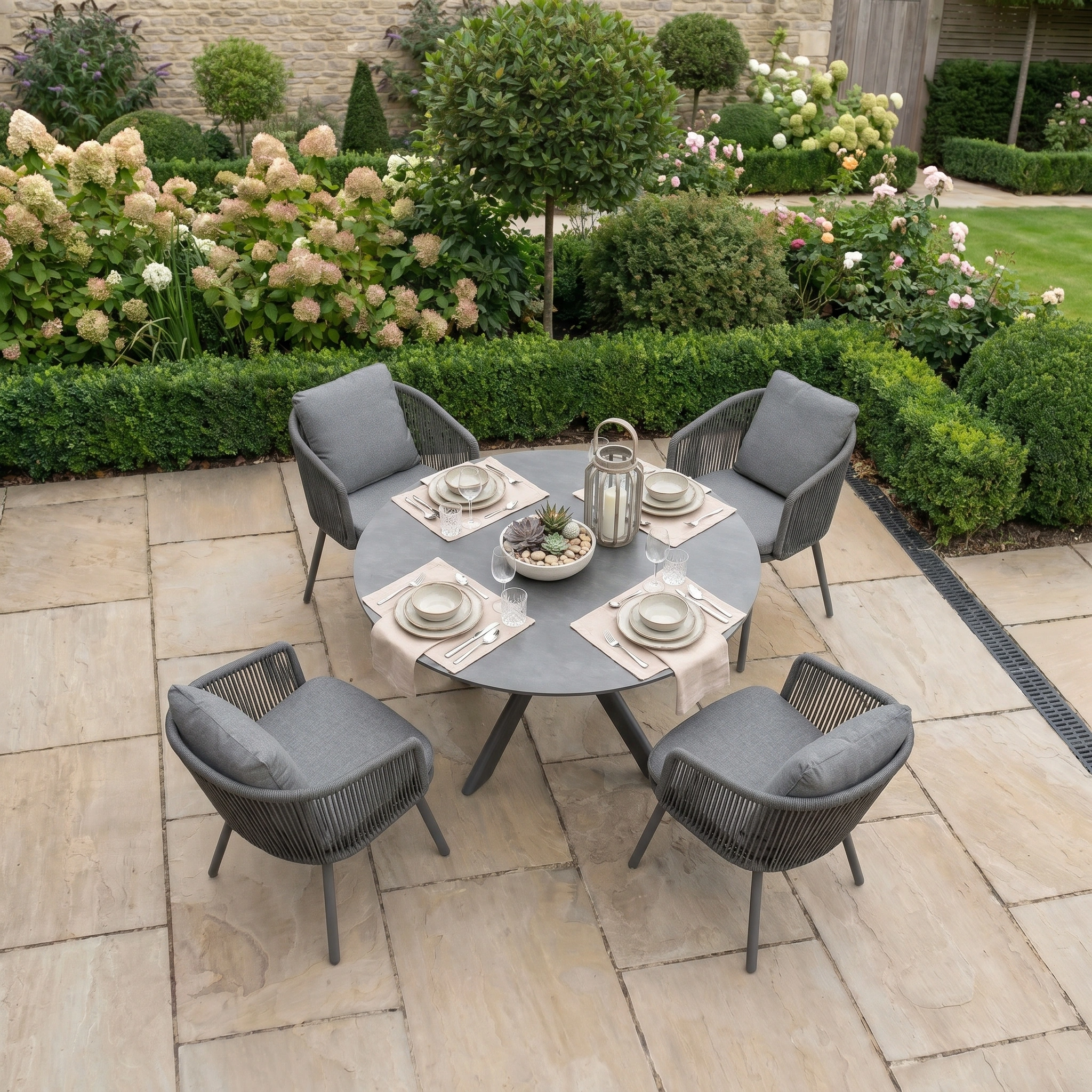 A Dekota 4 Seat Round Rope Dining Set with Albatross Table in Charcoal sits on a stone patio, surrounded by lush garden plants—perfect outdoor dining furniture for any garden retreat.