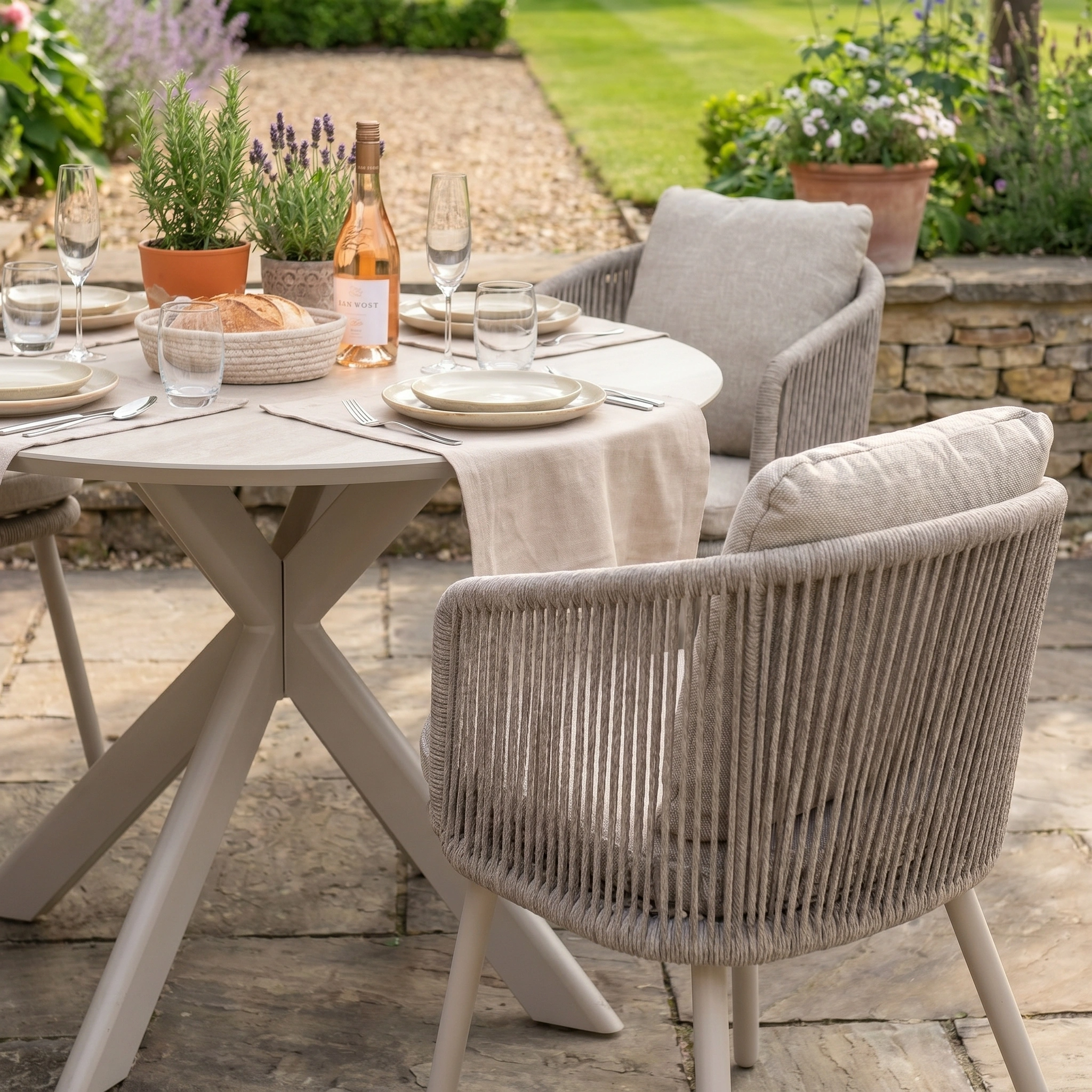 The Dekota 4 Seat Round Rope Dining Set with Albatross Table in Fawn features cushioned chairs on a stone patio, set for outdoor dining with wine, bread, plates, and glasses.