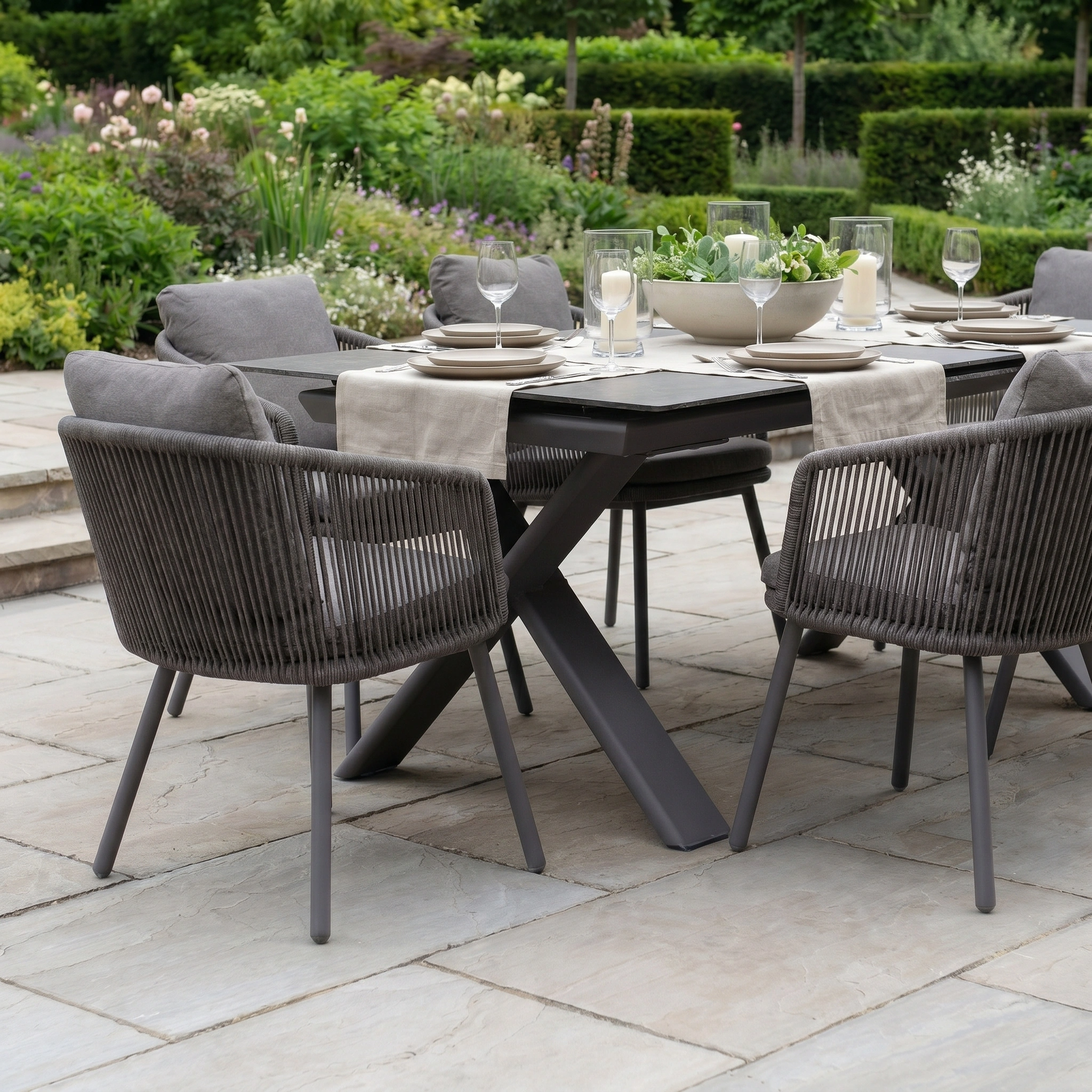 The Dekota 6 Seat Rope Dining Set with Albatross Extending Table in Charcoal features cushioned chairs, stylishly arranged on a stone patio amid a lush garden.