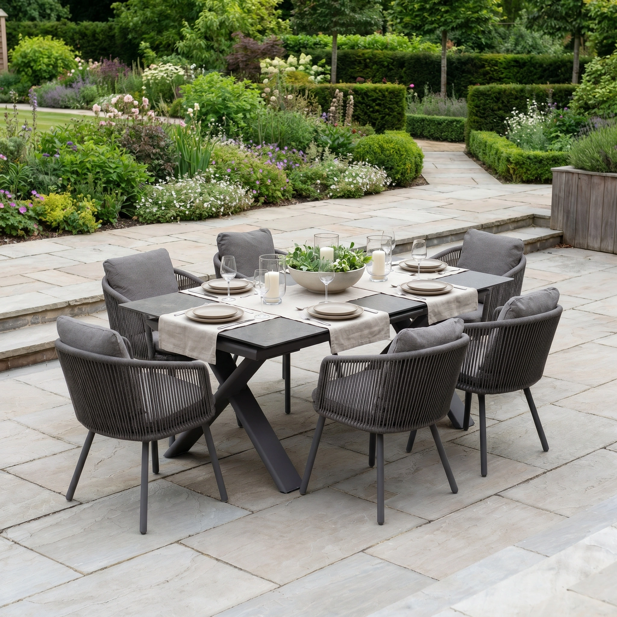 The Dekota 6 Seat Rope Dining Set with Albatross Extending Table in Charcoal seats six, featuring gray chairs, neutral tableware, and a picturesque garden backdrop for stylish outdoor dining.