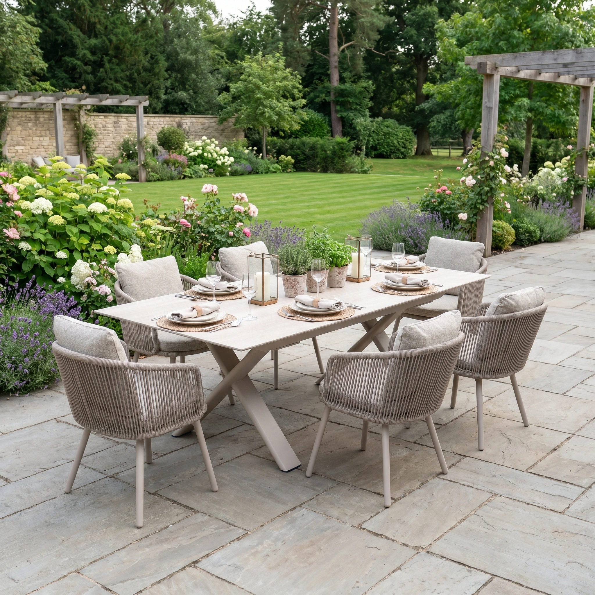 The Dekota 6 Seat Rope Dining Set with Albatross Extending Table in Fawn sits on the patio, surrounded by lush greenery and blooming flowers—ideal for outdoor dining in a tranquil garden setting.