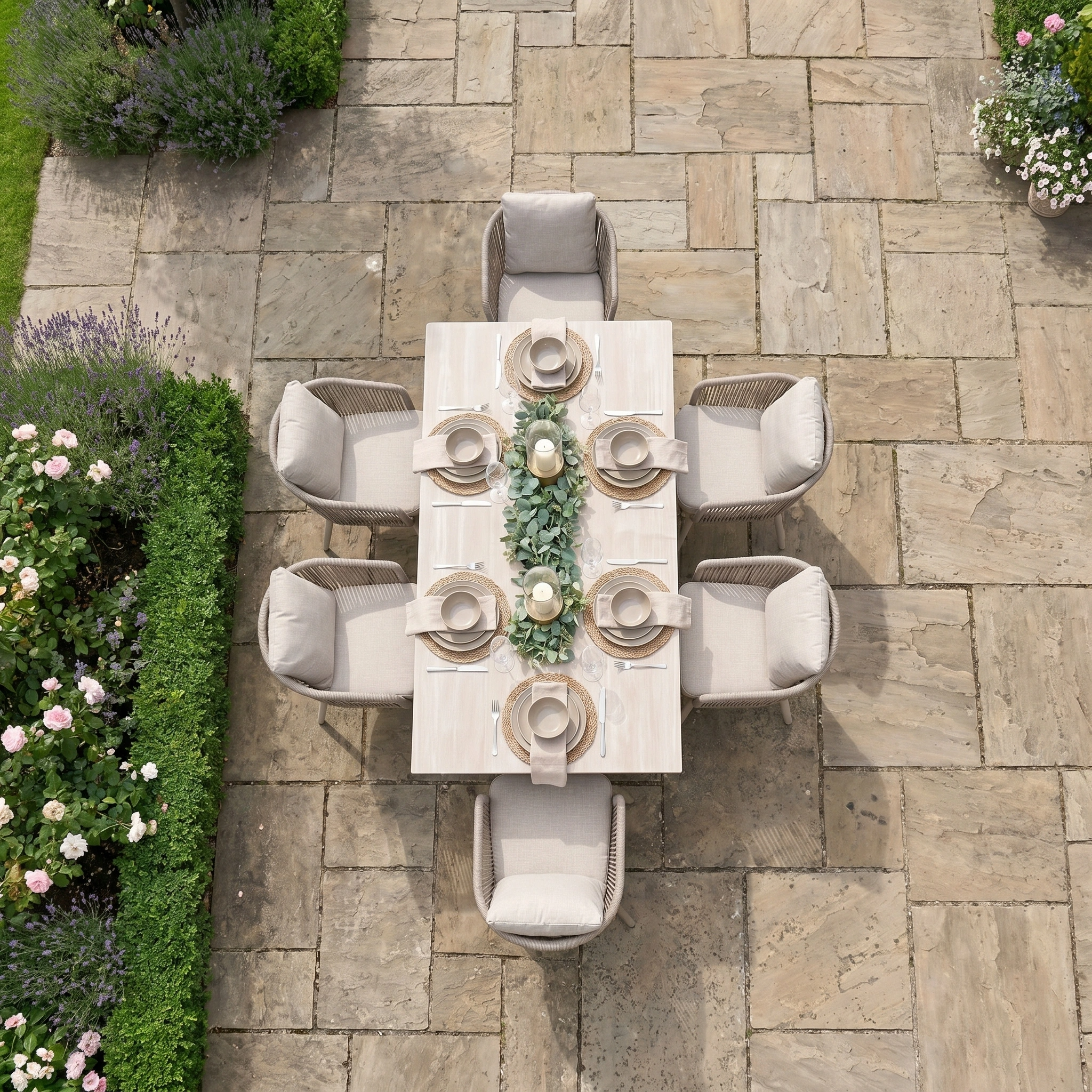 The Dekota 6 Seat Rope Dining Set with Albatross Extending Table in Fawn is perfect for outdoor meals on a stone patio, surrounded by lush greenery.