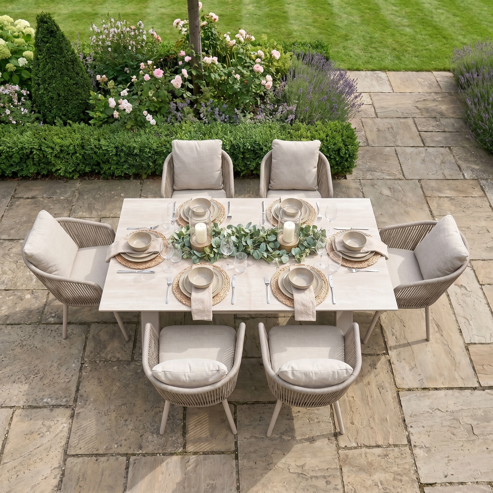 The Dekota 6 Seat Rope Dining Set with Albatross Extending Table in Fawn is shown on a stone patio with neutral decor, beside a lush garden and green grass.