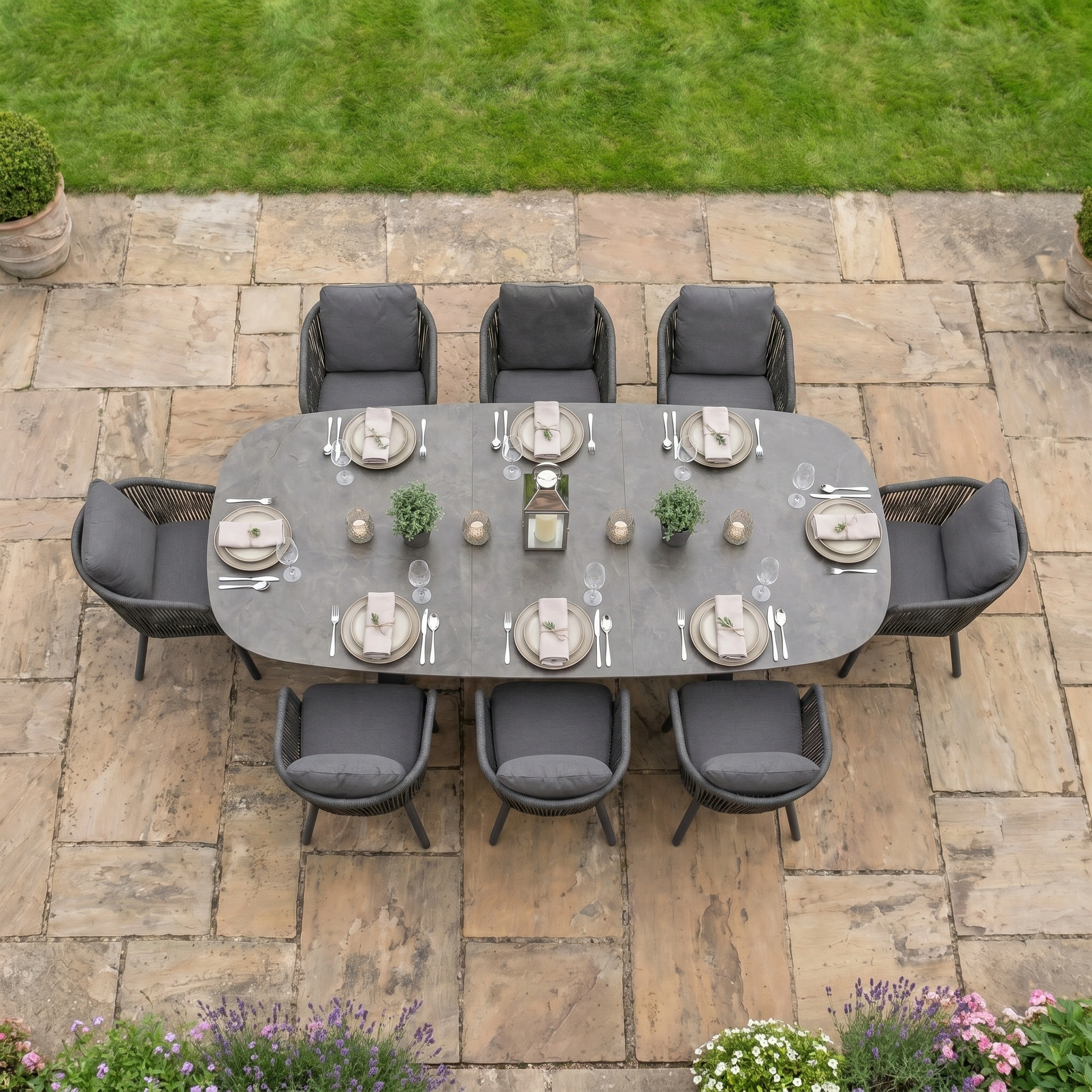 Dekota 8 Seat Oval Rope Dining Set with Albatross Extending Table in Charcoal, styled with gray chairs on a stone patio and overlooking a green lawn.