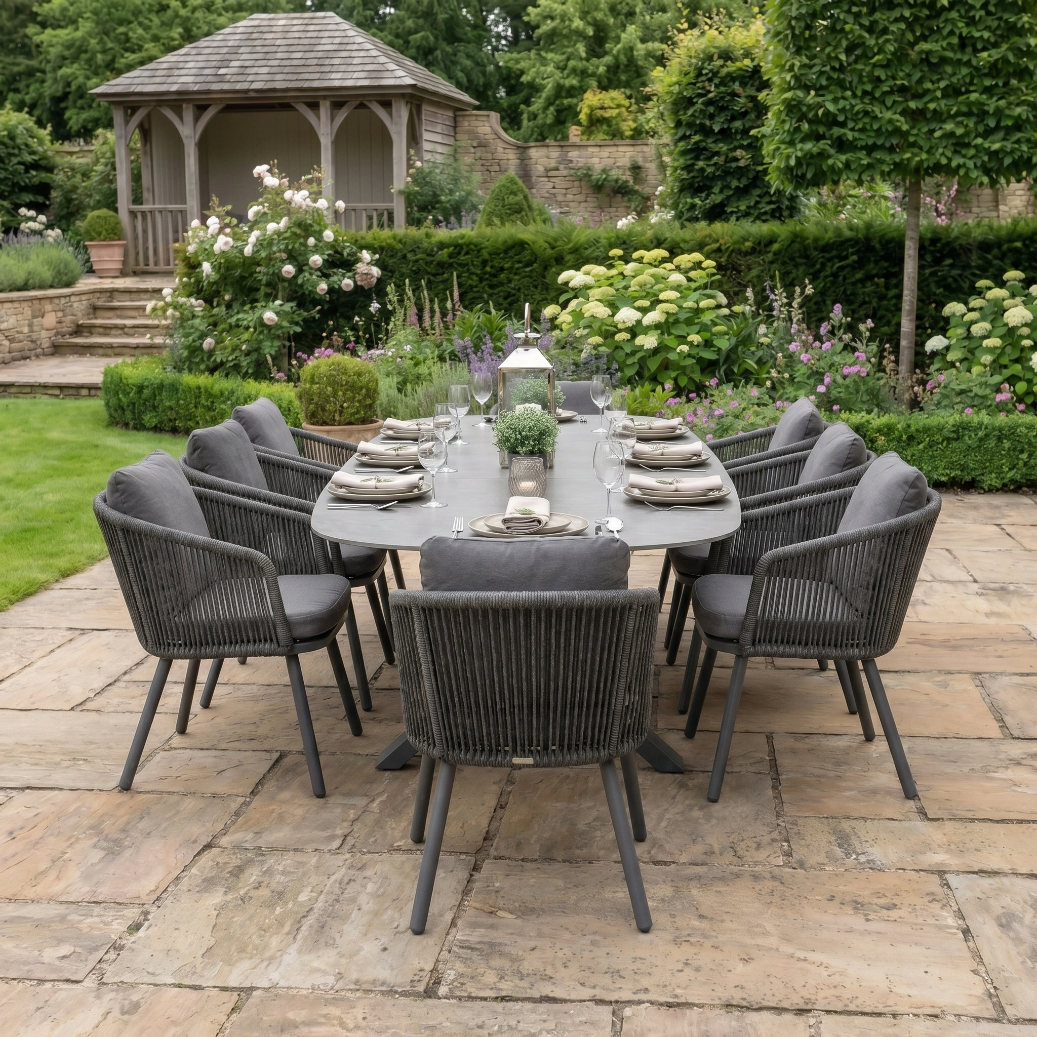 The Dekota 8 Seat Oval Rope Dining Set with Albatross Extending Table in Charcoal features a sleek sintered stone tabletop, set for outdoor dining amid a landscaped garden with a gazebo in the background.
