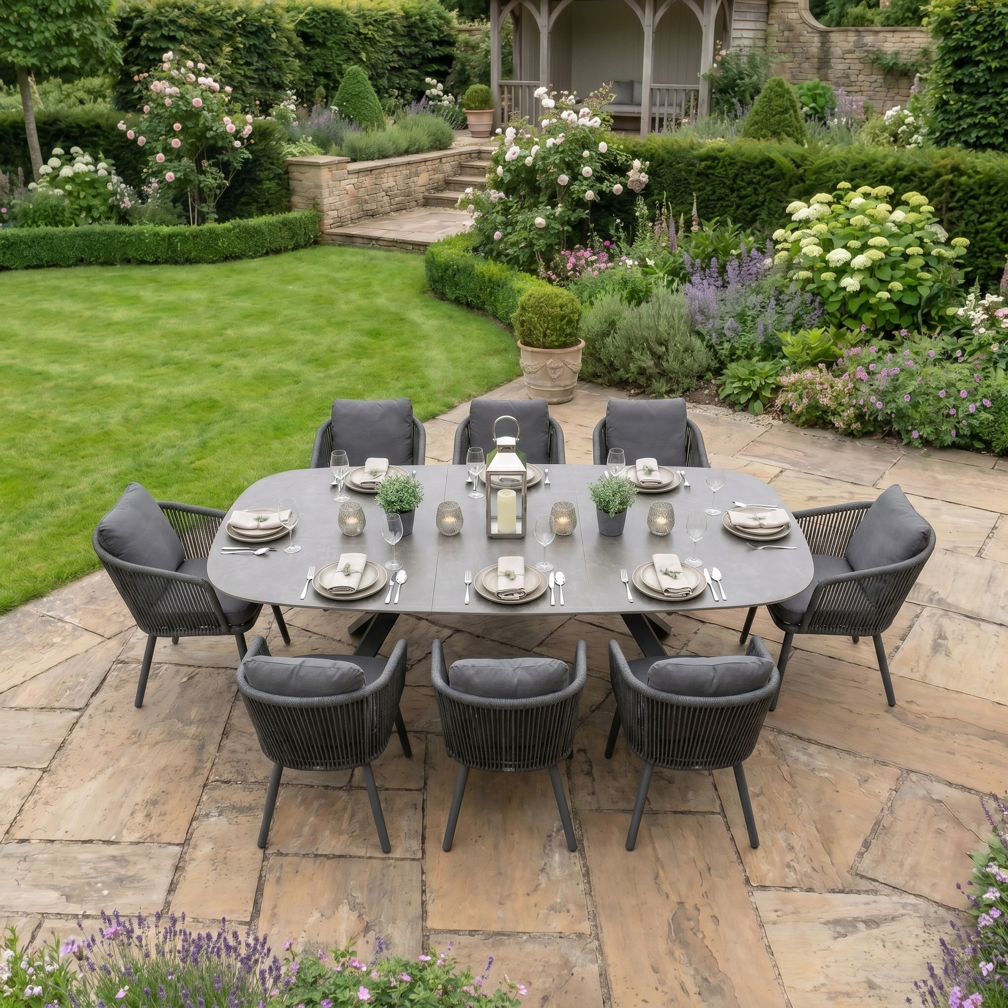 The Dekota 8 Seat Oval Rope Dining Set with Albatross Extending Table in Charcoal features a stylish, durable design perfect for outdoor dining, beautifully showcased on a stone patio among lush greenery and vibrant flowers.