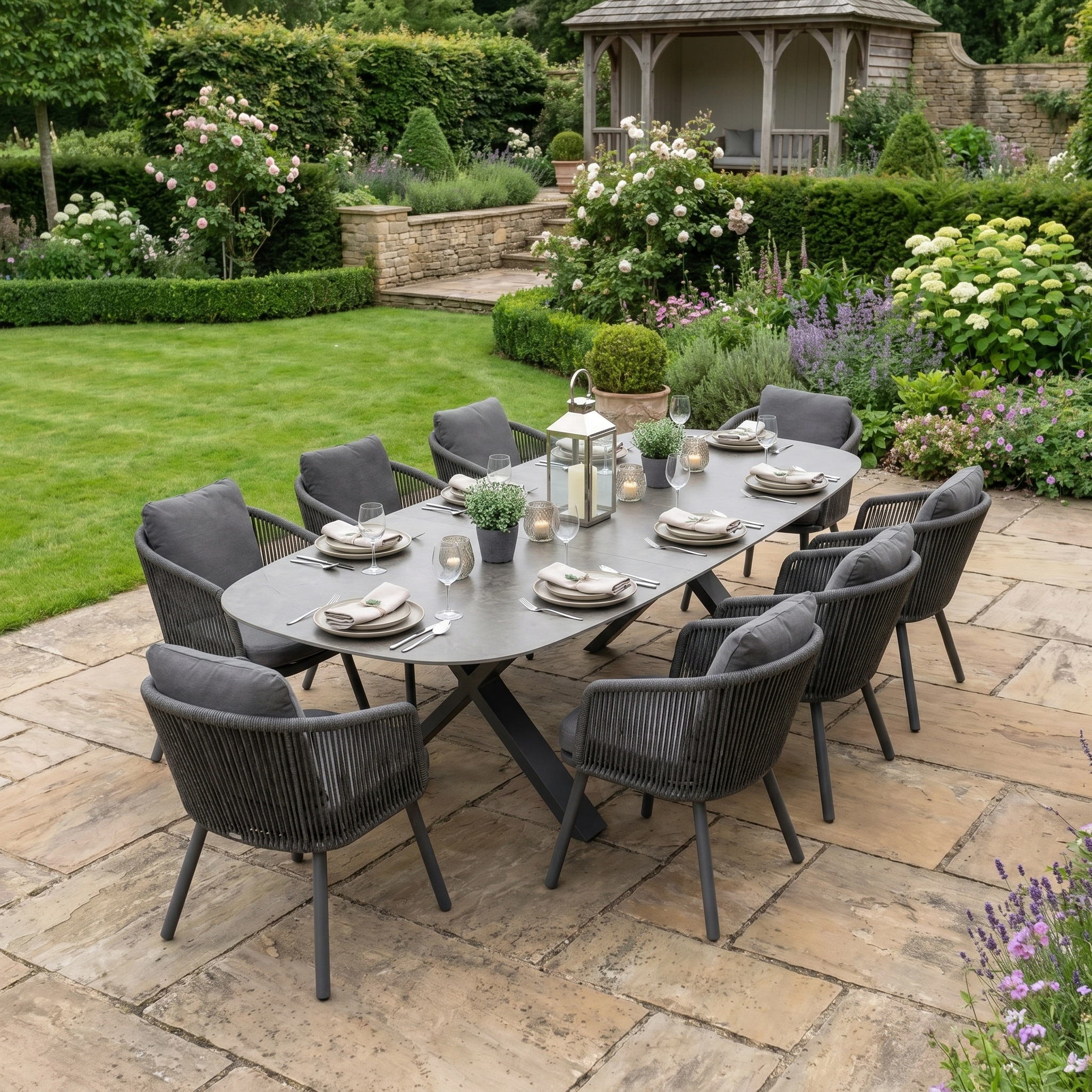 The Dekota 8 Seat Oval Rope Dining Set with Albatross Extending Table in Charcoal features an elegant oval dining table and eight chairs, perfectly arranged on a patio in a lush garden.