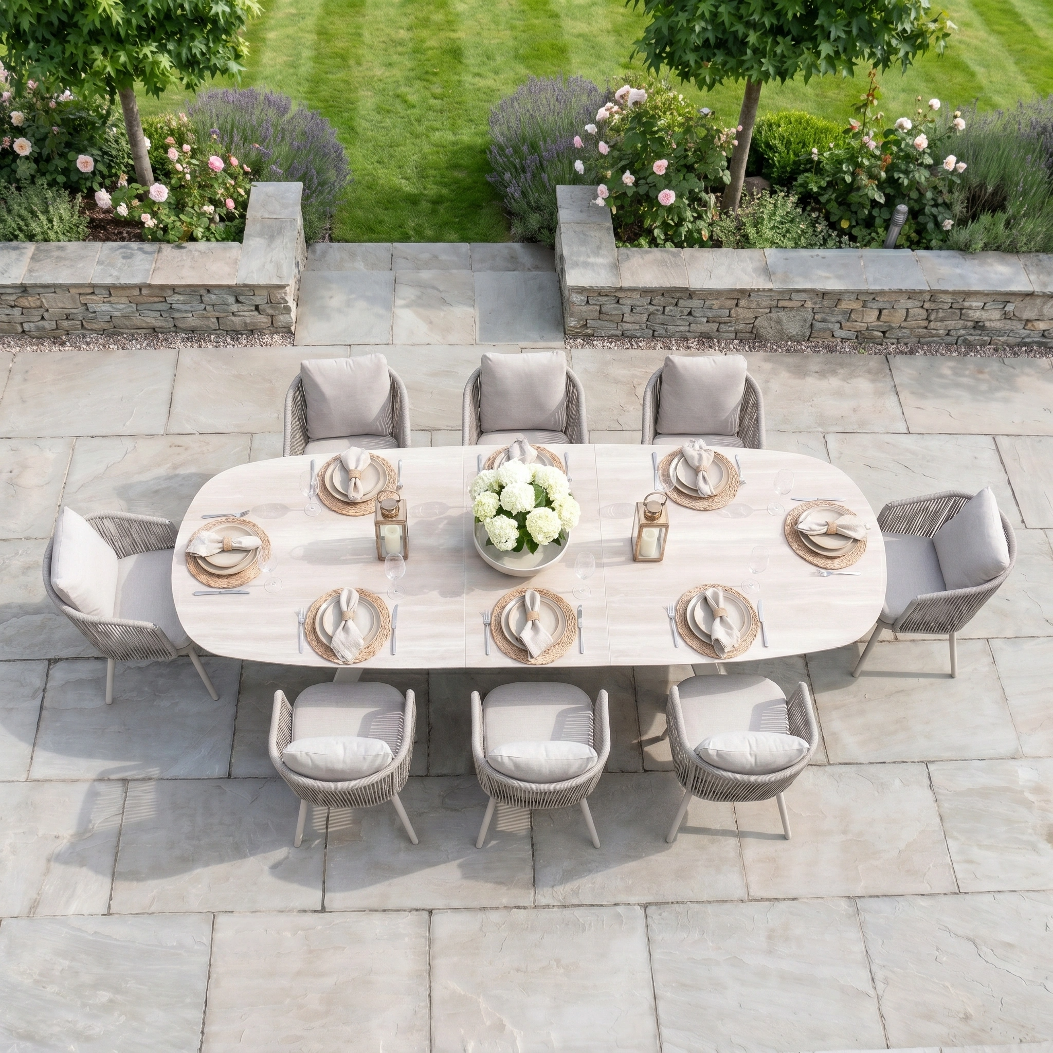 The Dekota 8 Seat Oval Rope Dining Set with Albatross Extending Table in Fawn features woven chairs and a light wood extending table, perfect for gatherings on a stone patio with a lush garden backdrop.