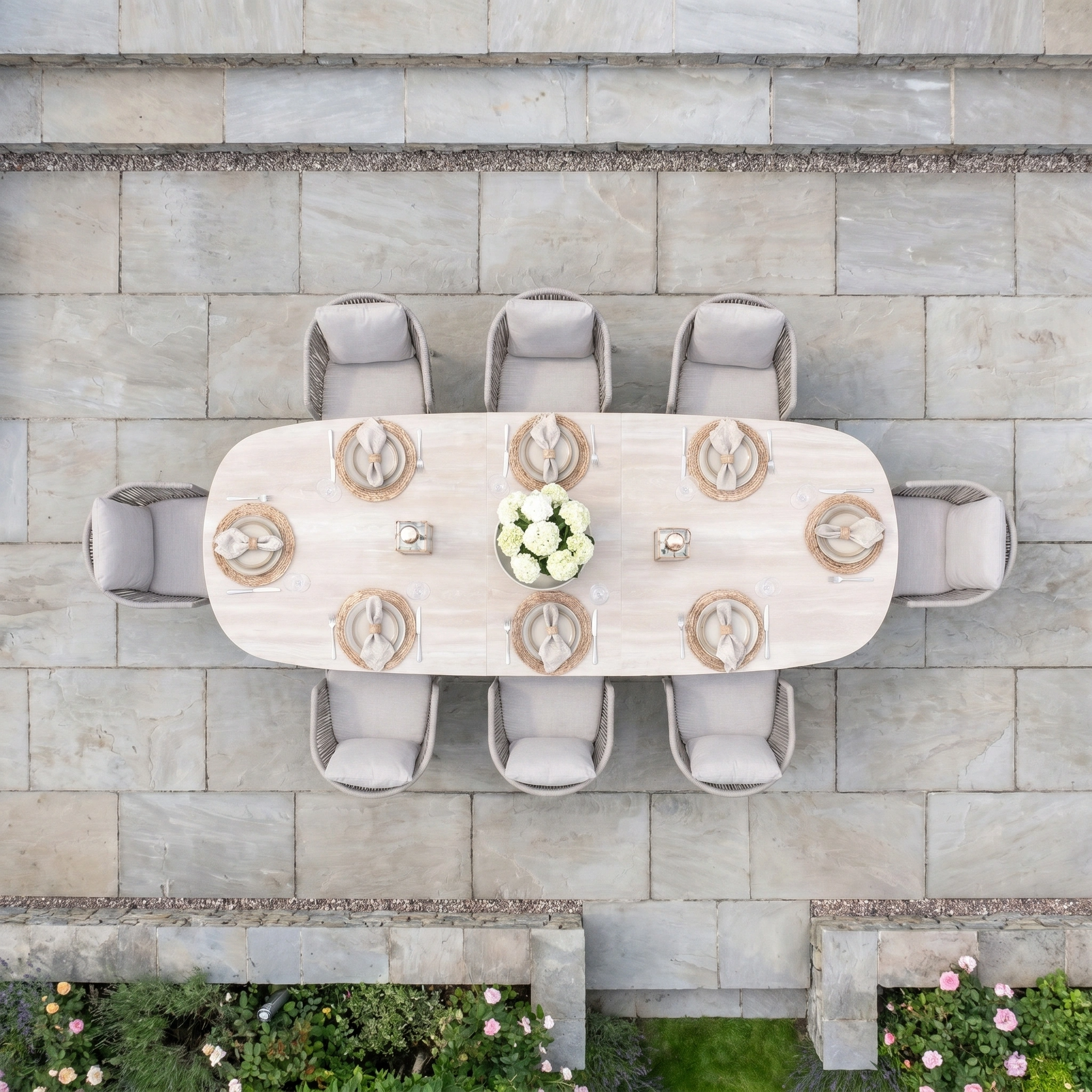 Aerial view of the Dekota 8 Seat Oval Rope Dining Set with Albatross Extending Table in Fawn, elegantly arranged for eight with plates, napkins, and a flower centerpiece on stone tiles—ideal for outdoor gatherings.