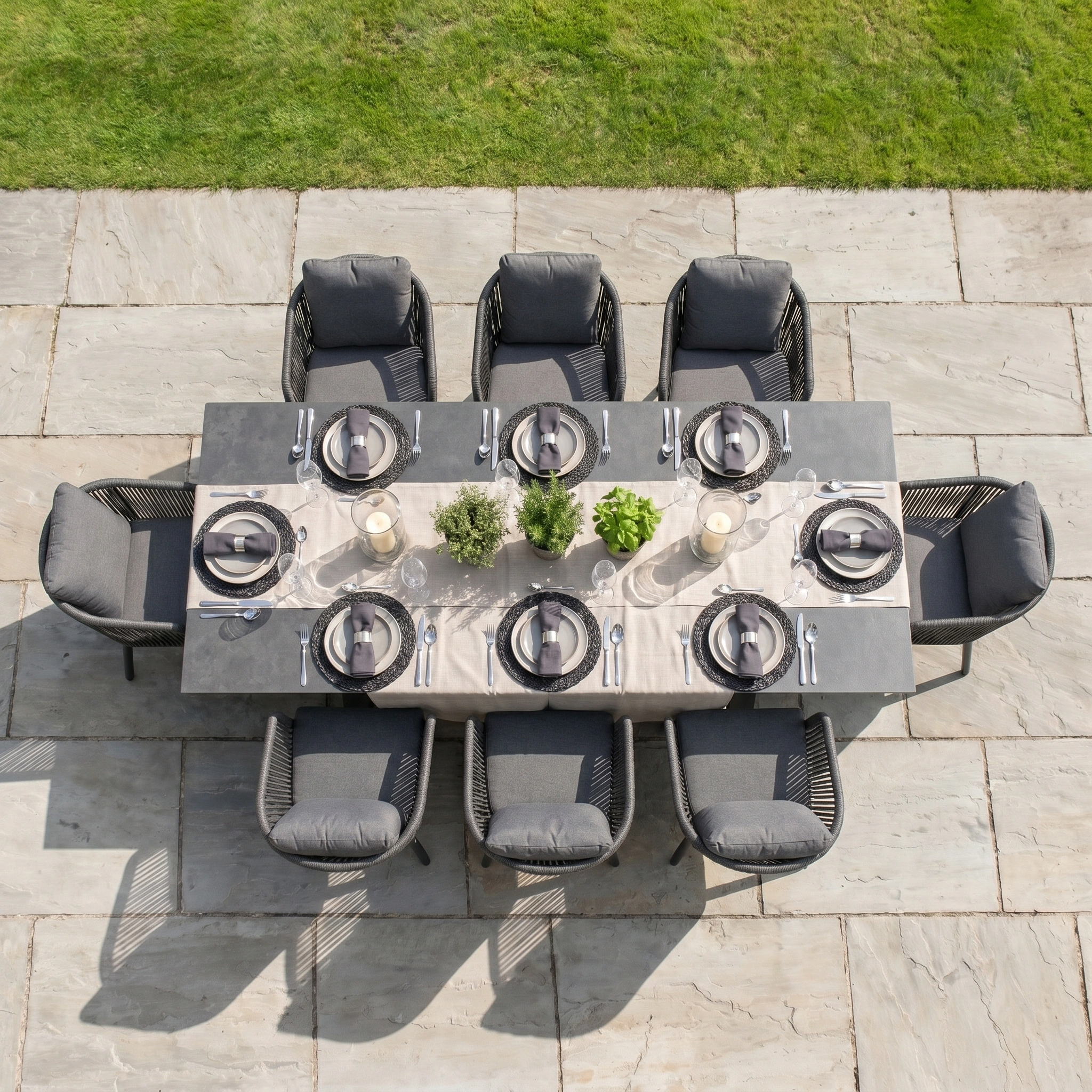 The Dekota 8 Seat Rope Dining Set with Albatross Extending Table in Charcoal is arranged on a stone patio with plants as centerpieces—stylish and perfect for outdoor entertaining beside the grass.