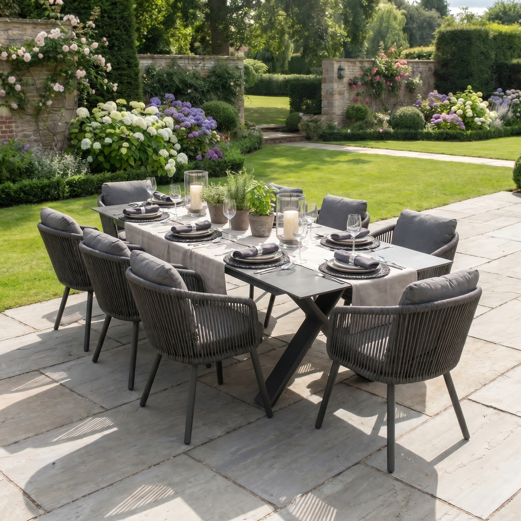 The Dekota 8 Seat Rope Dining Set with Albatross Extending Table in Charcoal is perfect for stylish outdoor dining on a stone patio in a lush, blooming garden.