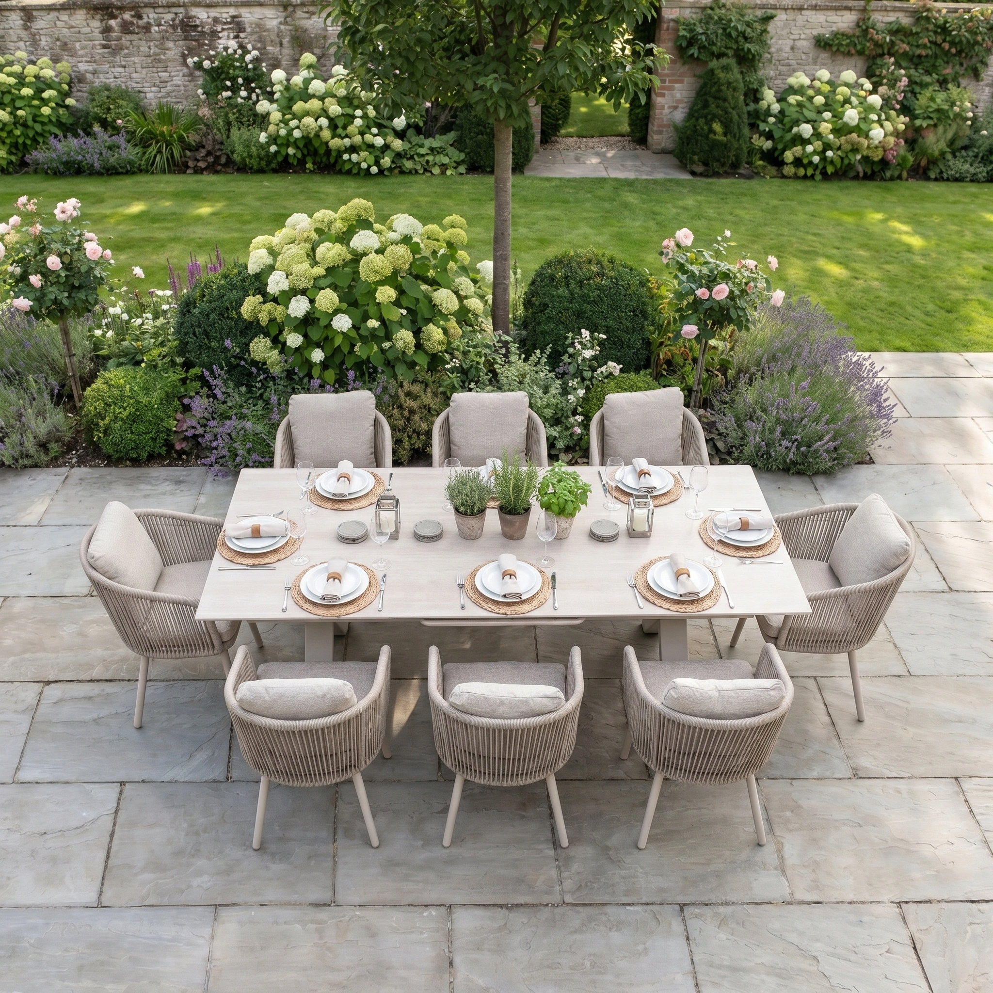The Dekota 8 Seat Rope Dining Set with Albatross Extending Table in Fawn is scratch and UV-resistant outdoor furniture, perfect for entertaining on a stone patio amid lush gardens and blooming flowers.