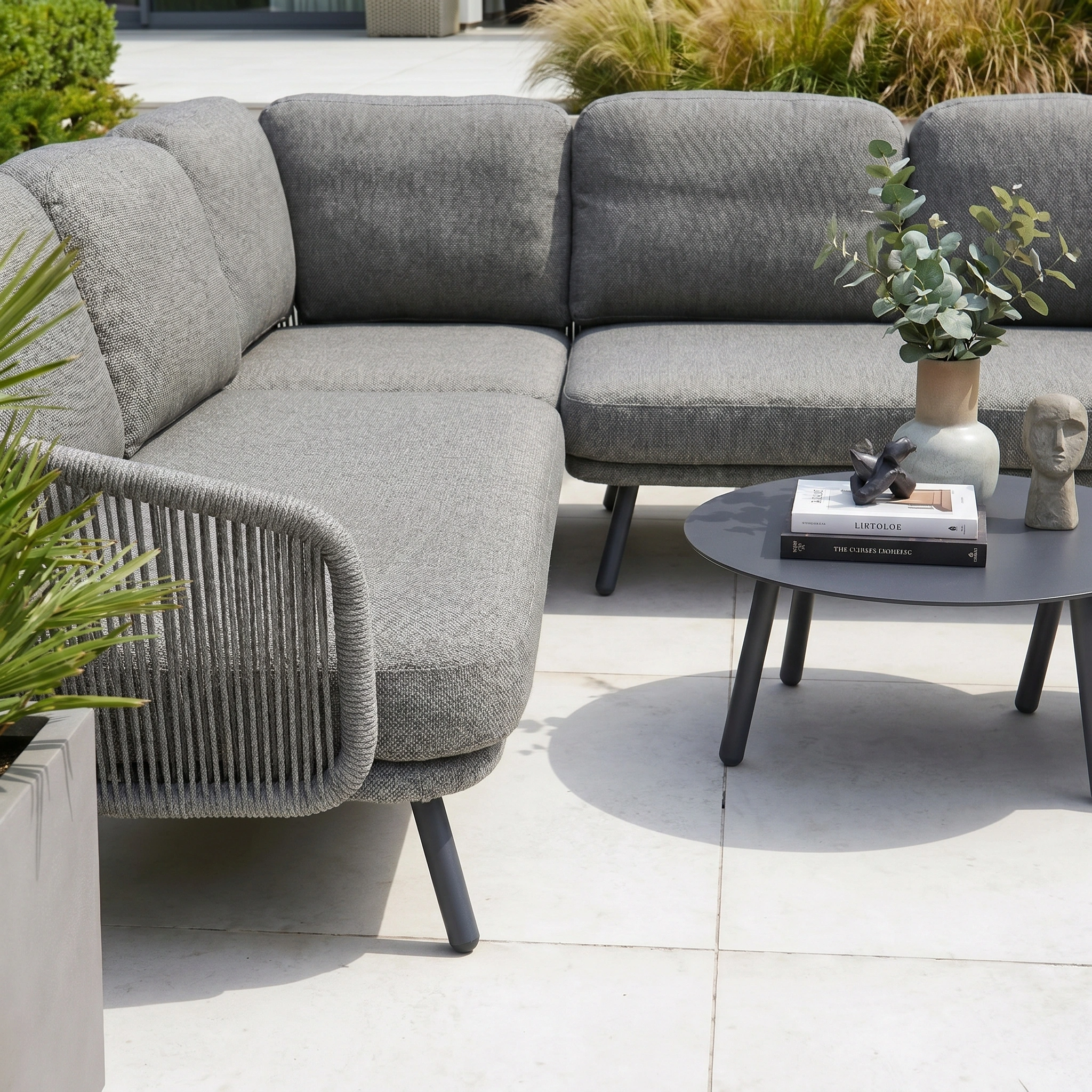 The Dekota Corner Sofa Set in Charcoal, paired with a round black table and plants, transforms this white patio—outdoor furniture designed for comfort and modern style.