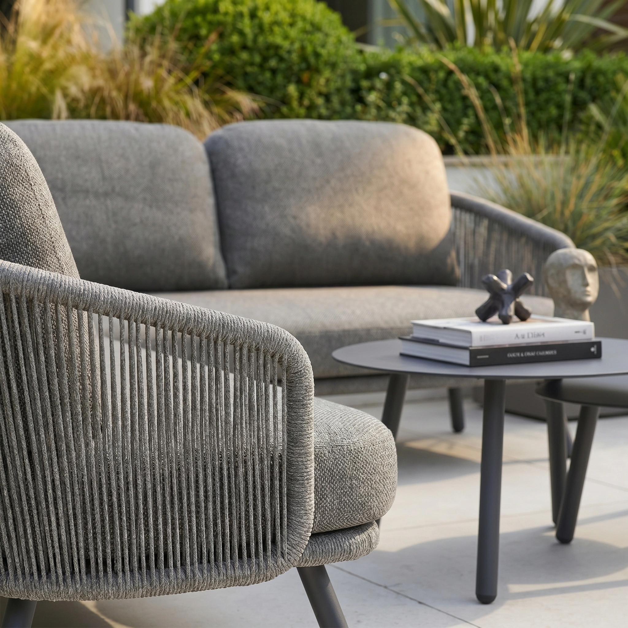 Modern outdoor patio with gray cushioned seating from the Dekota Corner Sofa Set in Charcoal, a round table, and books, surrounded by green plants—perfect for relaxing with stylish outdoor furniture.