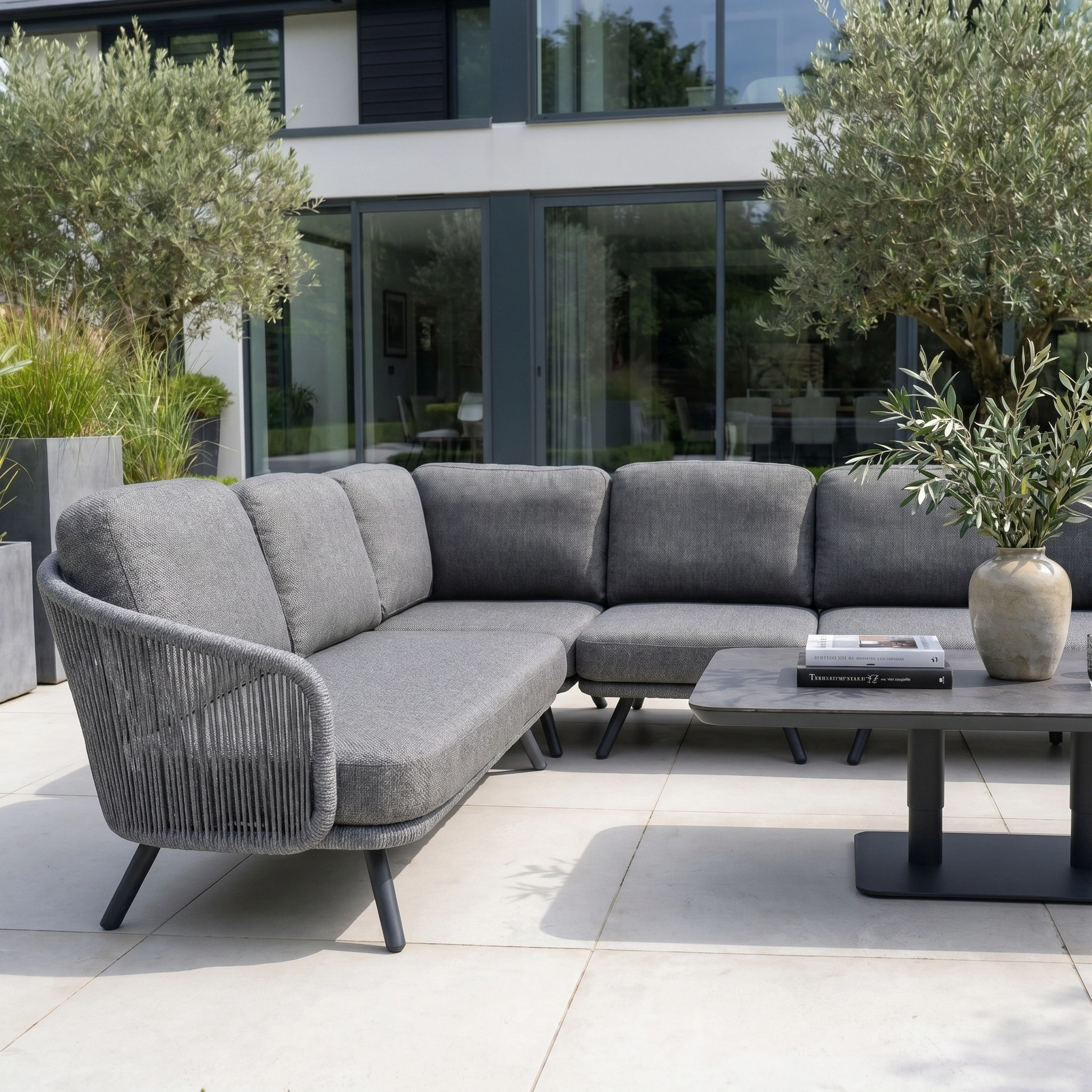 The Dekota Large Corner Sofa Dining Set with Altura Rising Table in Charcoal sits on a patio with potted plants, showcasing a hydrophobic rope weave and large glass doors in the background.