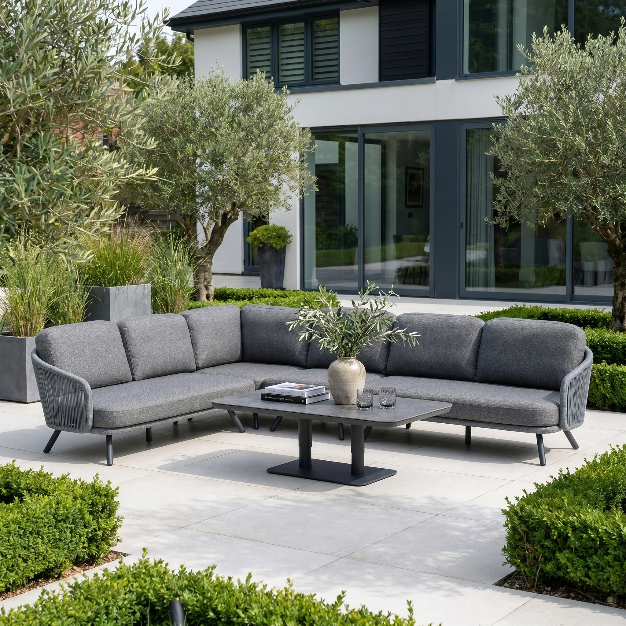Dekota Large Corner Sofa Dining Set with Altura Rising Table in Charcoal on a patio, surrounded by olive trees and manicured hedges.