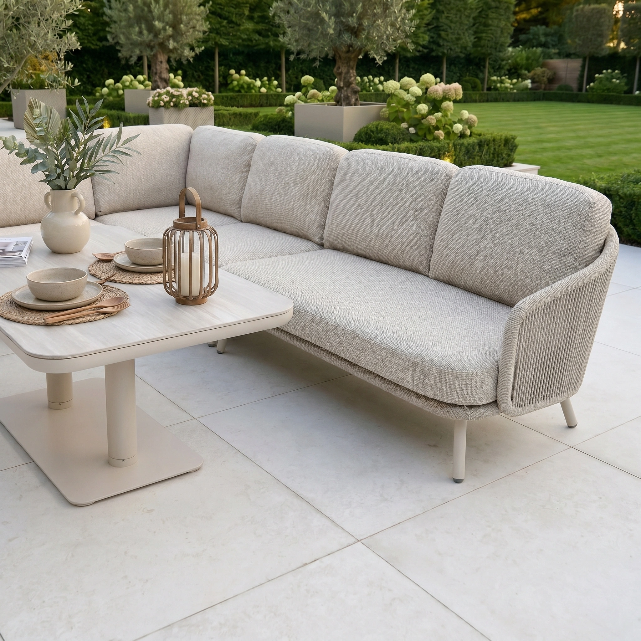 The Dekota Large Corner Sofa Dining Set with Altura Rising Table in Fawn sits on a tiled patio with decor, offering an inviting space to relax or entertain outdoors against a garden backdrop.