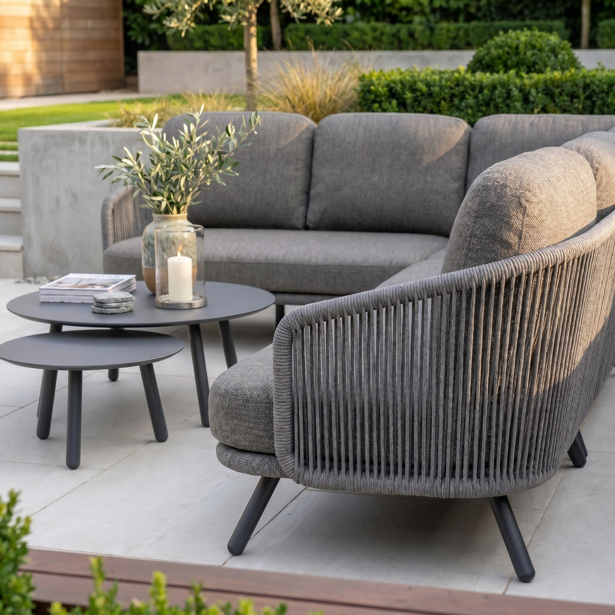 The Dekota Large Corner Sofa Set in Charcoal, paired with round tables topped with candles and a vase, creates an inviting patio surrounded by lush greenery.