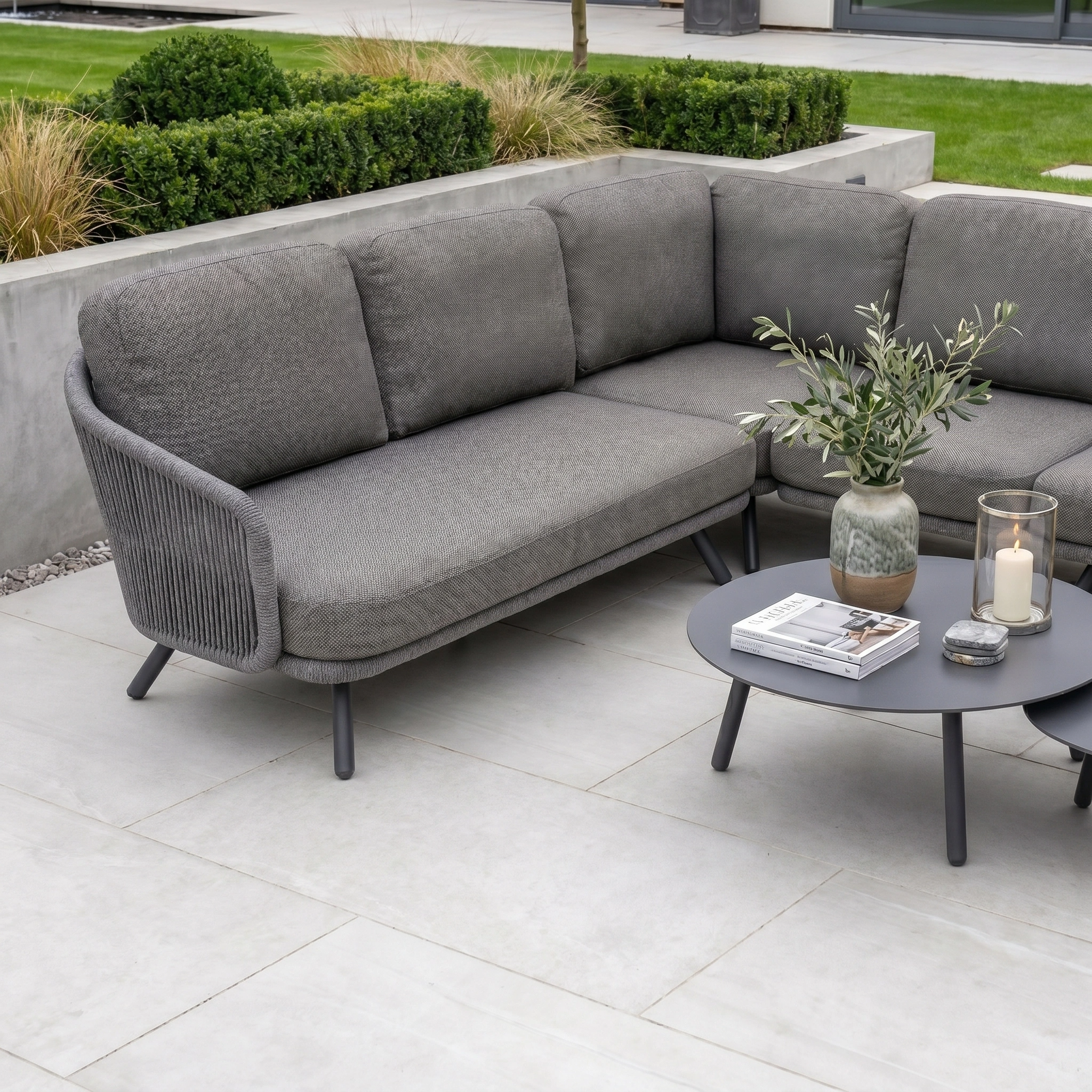 The Dekota Large Corner Sofa Set in Charcoal creates a modern outdoor space, paired with a sleek round black coffee table on a tiled patio and surrounded by lush plants.