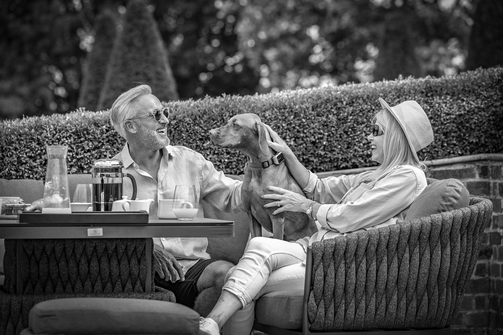 A golden retriever lounges on a light gray outdoor couch with a patterned pillow, near a modern coffee table with cups. The Bay Large Corner Group Set with Aluminium Frame in Latte features a powder-coated aluminium frame and durable Olefin material, ensuring both style and resilience in any weather.