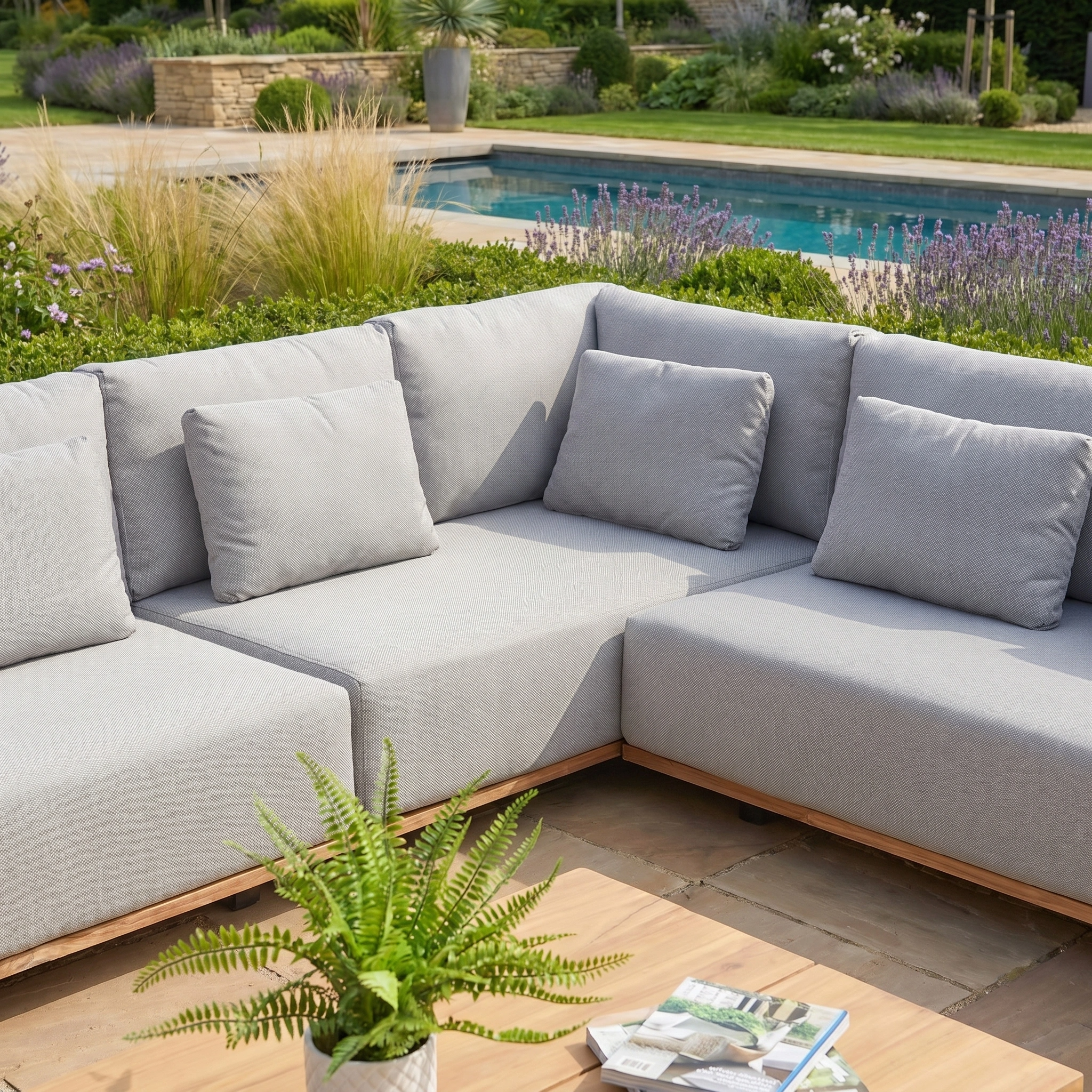 The Eternity Right Hand Teak Corner Group in Light Grey is a modular outdoor sectional sofa with cushions, placed beside a pool and complemented by surrounding plants and a wooden table.