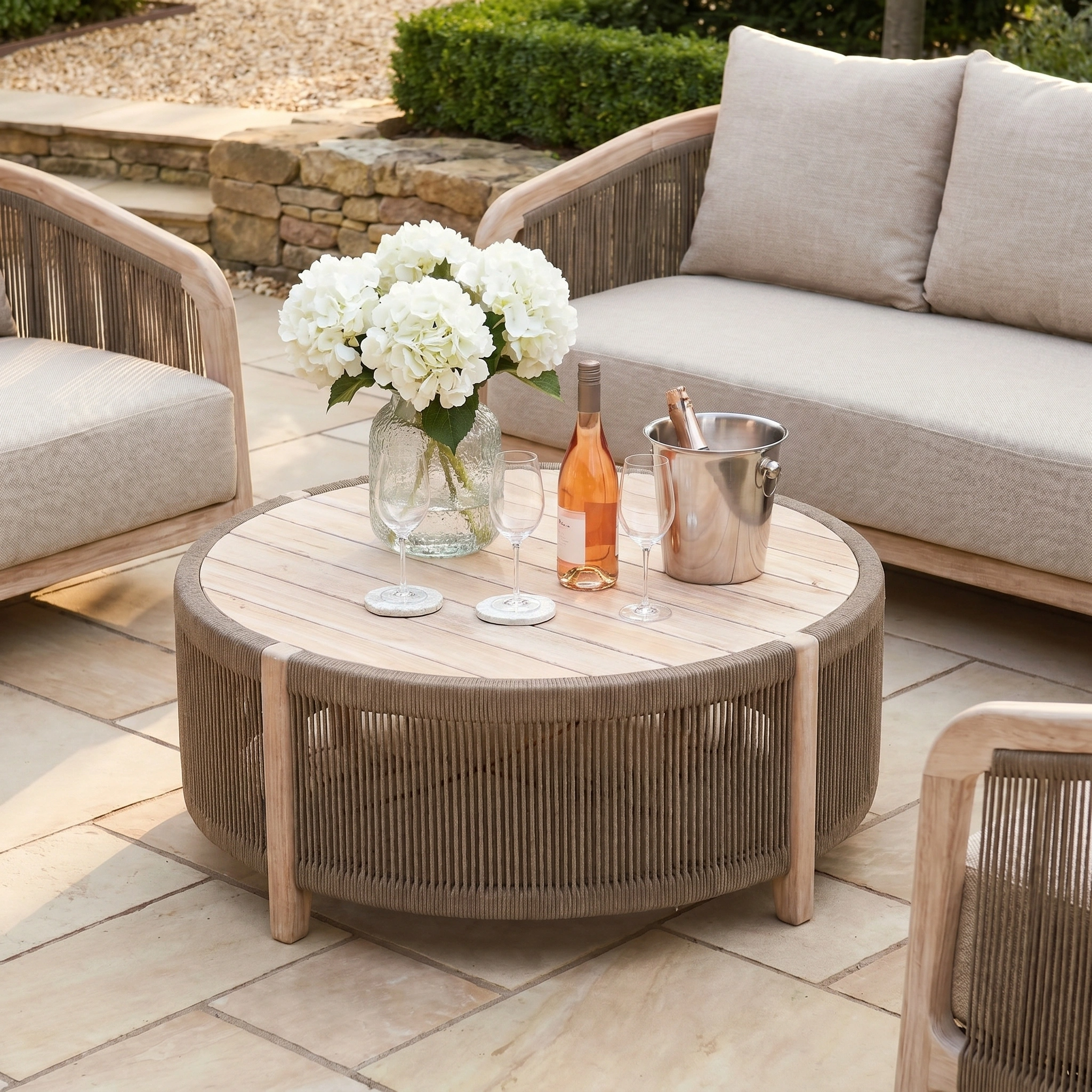 A round wooden patio table with flowers, wine, and glasses is paired with the Evora 3 Seat Sofa Set in Mocha for a stylish alfresco gathering.