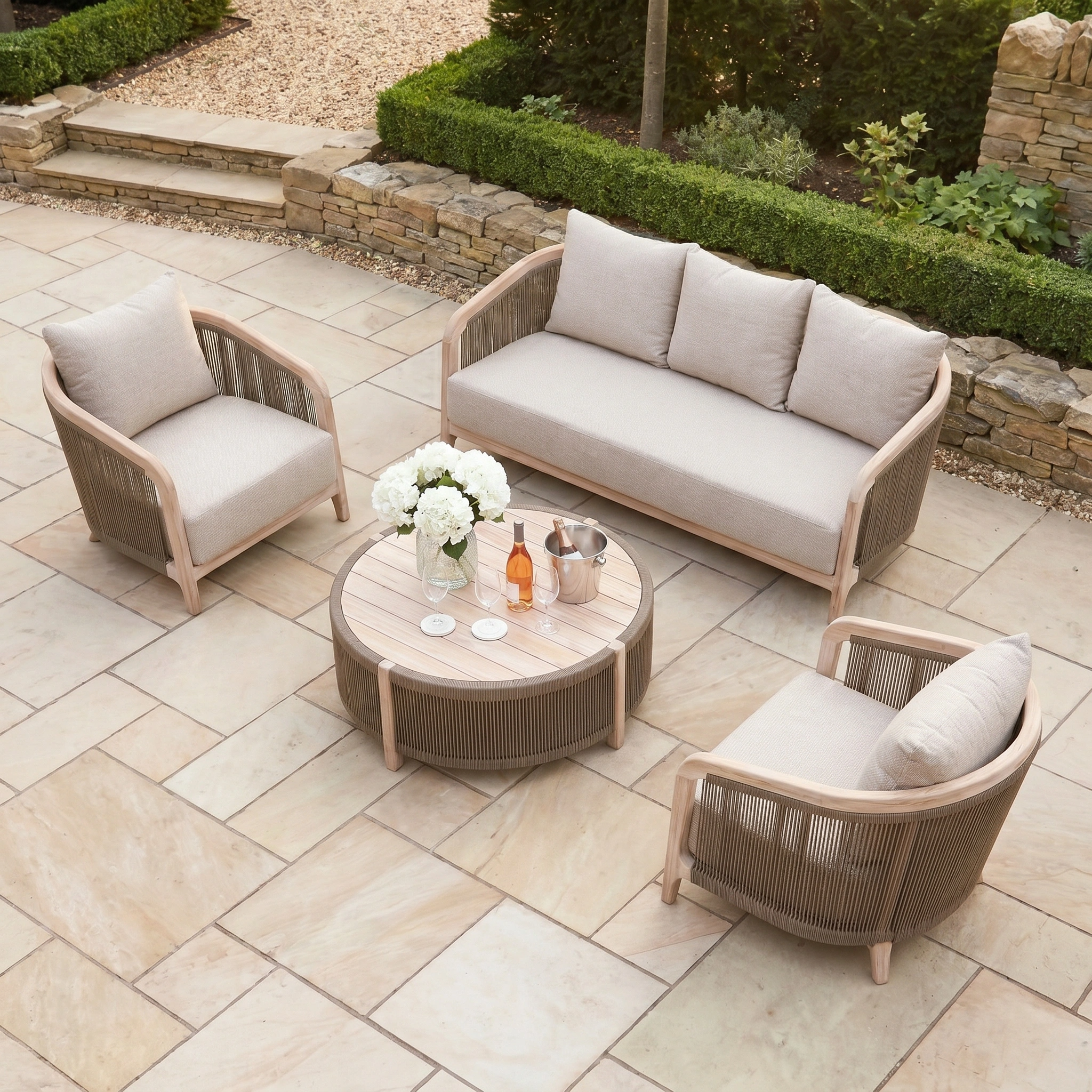 Evora 3 Seat Sofa Set in Mocha: Outdoor patio set includes a mocha teak sofa, two chairs, and a round table on a stone-tiled terrace.