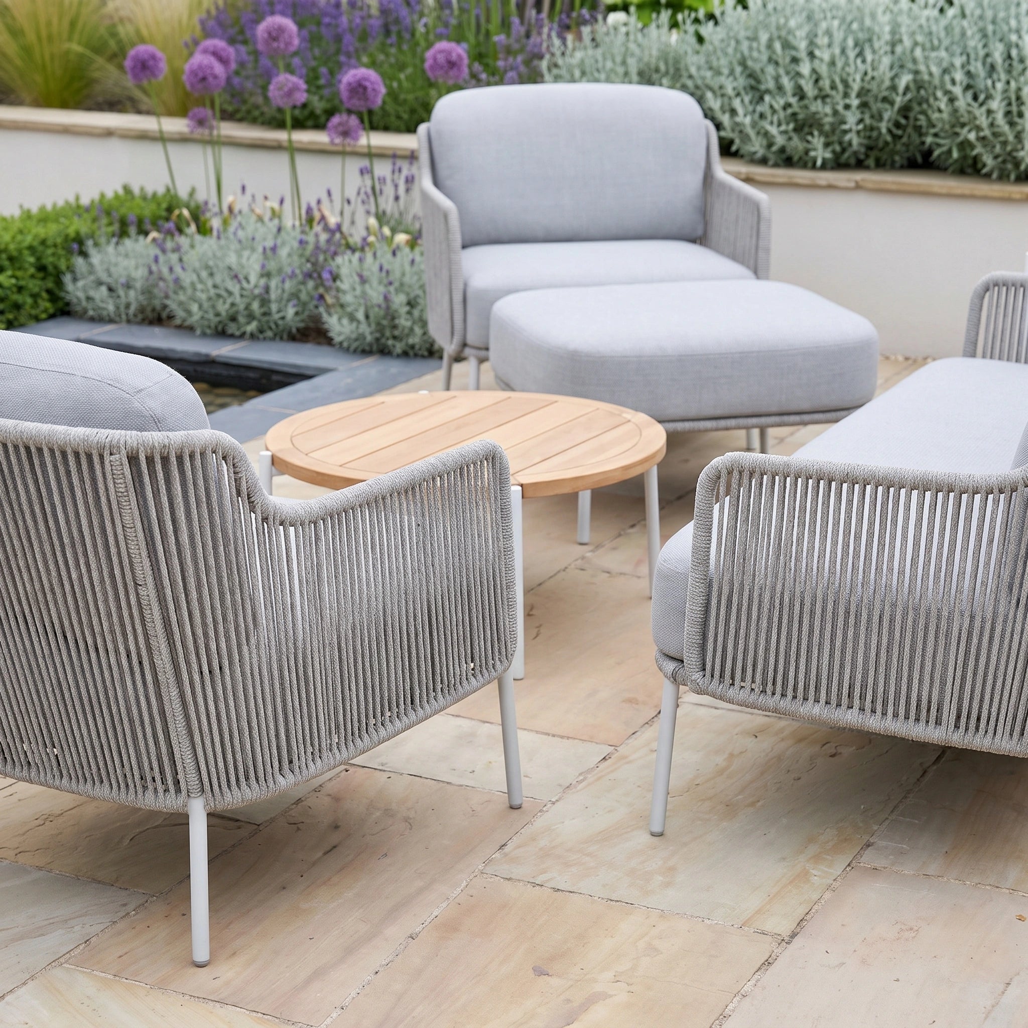 Four light grey Bernini 2 Seat Sofa Set pieces with teak tables and a matching footstool are arranged on stone tiles, creating a stylish retreat with the refined look of contemporary rope furniture.