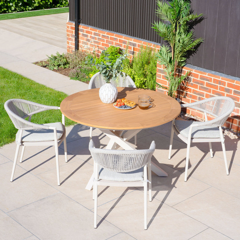 The Cloverly 4 Seat Round Dining Set with a Teak Table Top in Latte, including four woven chairs, is arranged with a vase and snacks on a sunlit paved outdoor area.