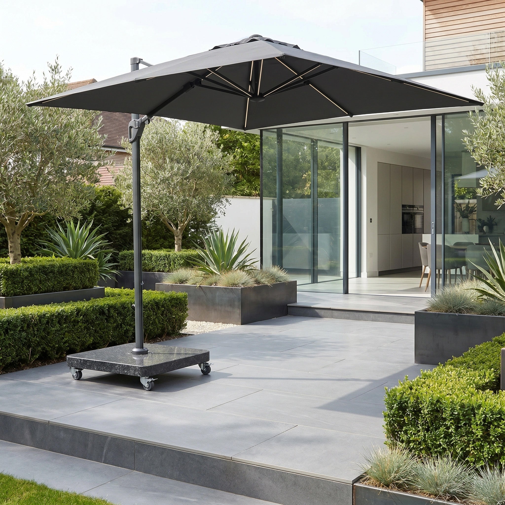 Modern patio featuring the Glow Challenger T2 3m Square Cantilever Parasol in Anthracite, manicured plants, and glass doors leading to a contemporary home.
