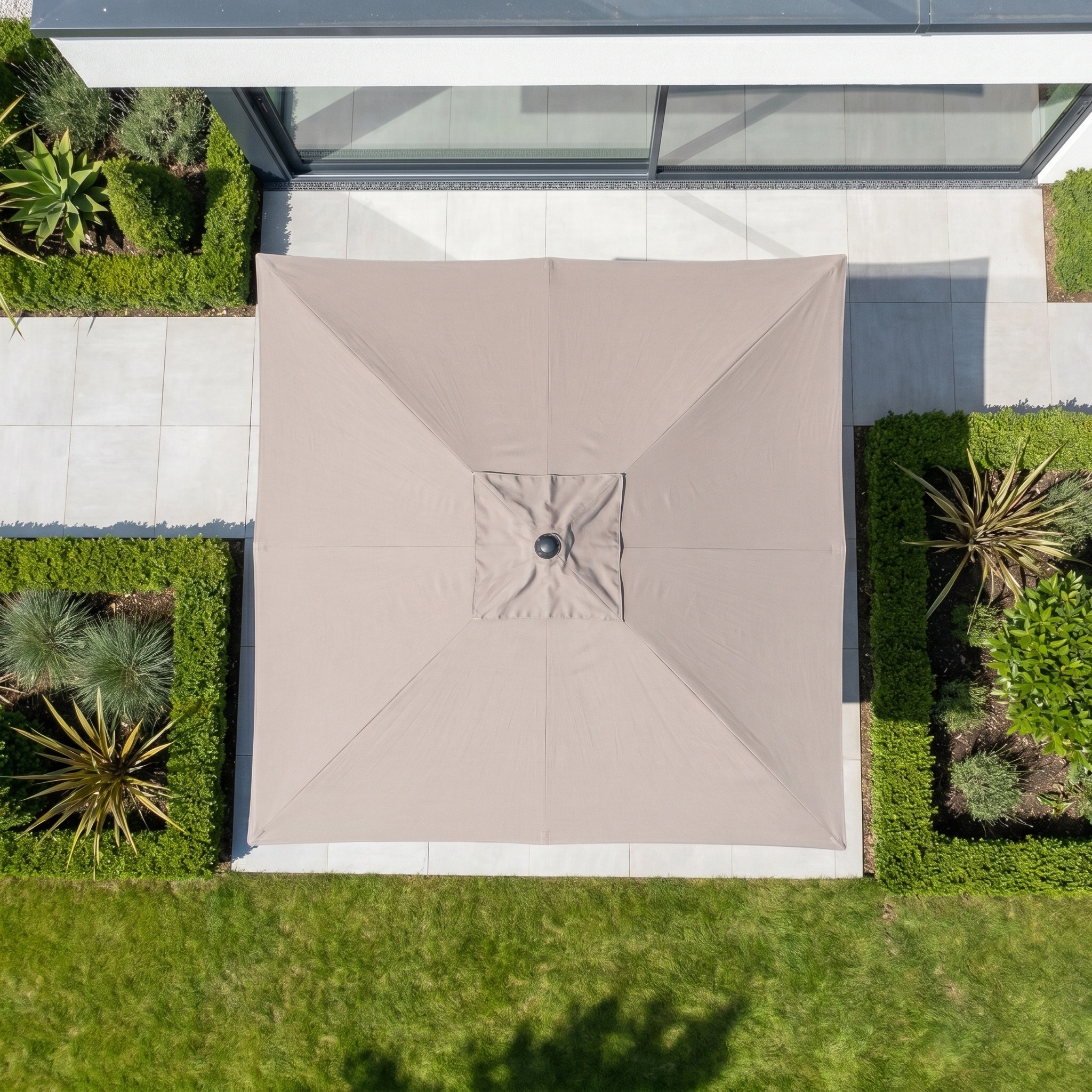 The Glow Challenger T2 3m Square Cantilever Parasol in Taupe, featuring a 90kg granite base, is pictured from above surrounded by green hedges and plants near a modern building.