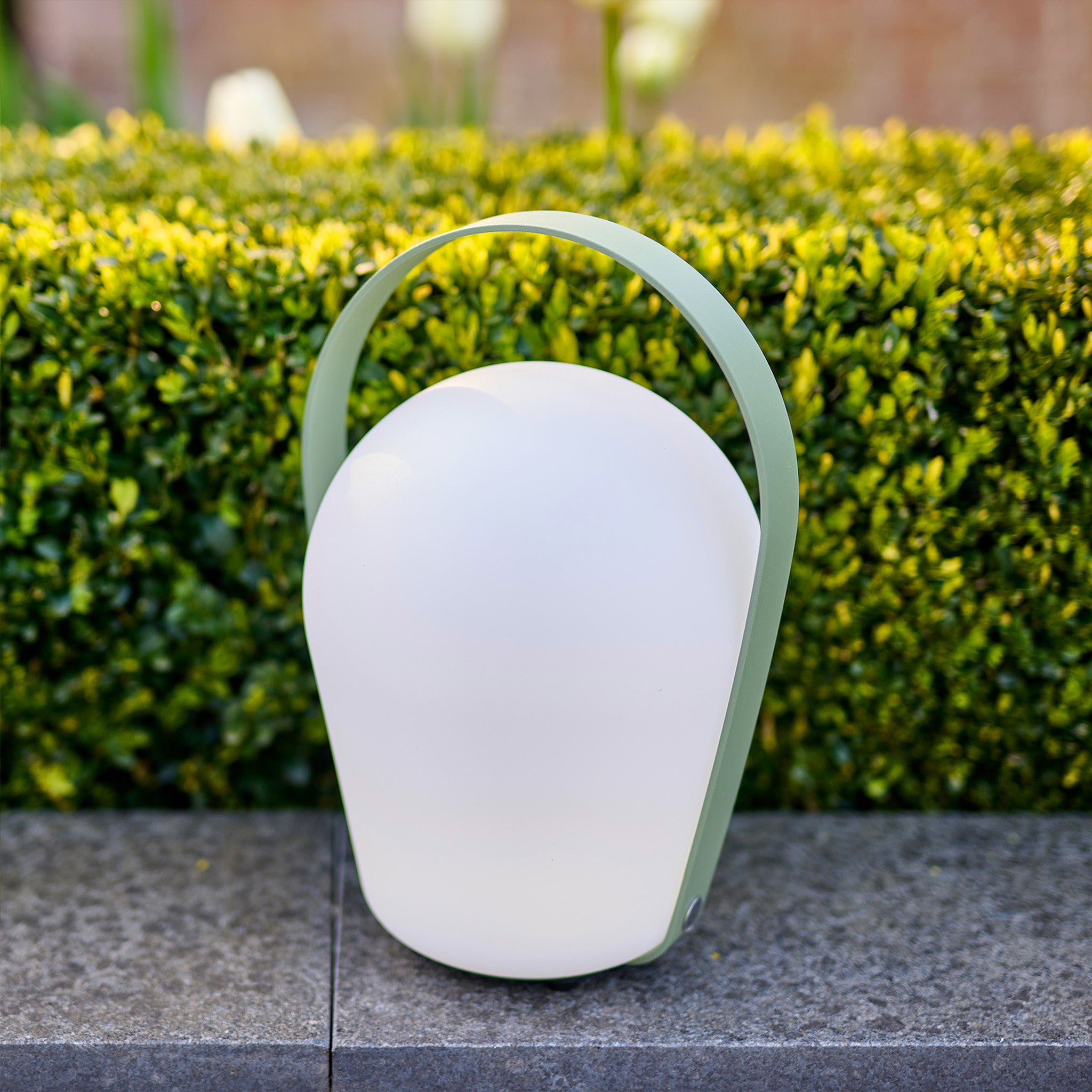 Introducing the Bob Large Table Light in Sage Green, situated on a stone surface and surrounded by lush green foliage. This portable garden lamp features a rechargeable function for your convenience and is weather-resistant, perfect for all your outdoor adventures.