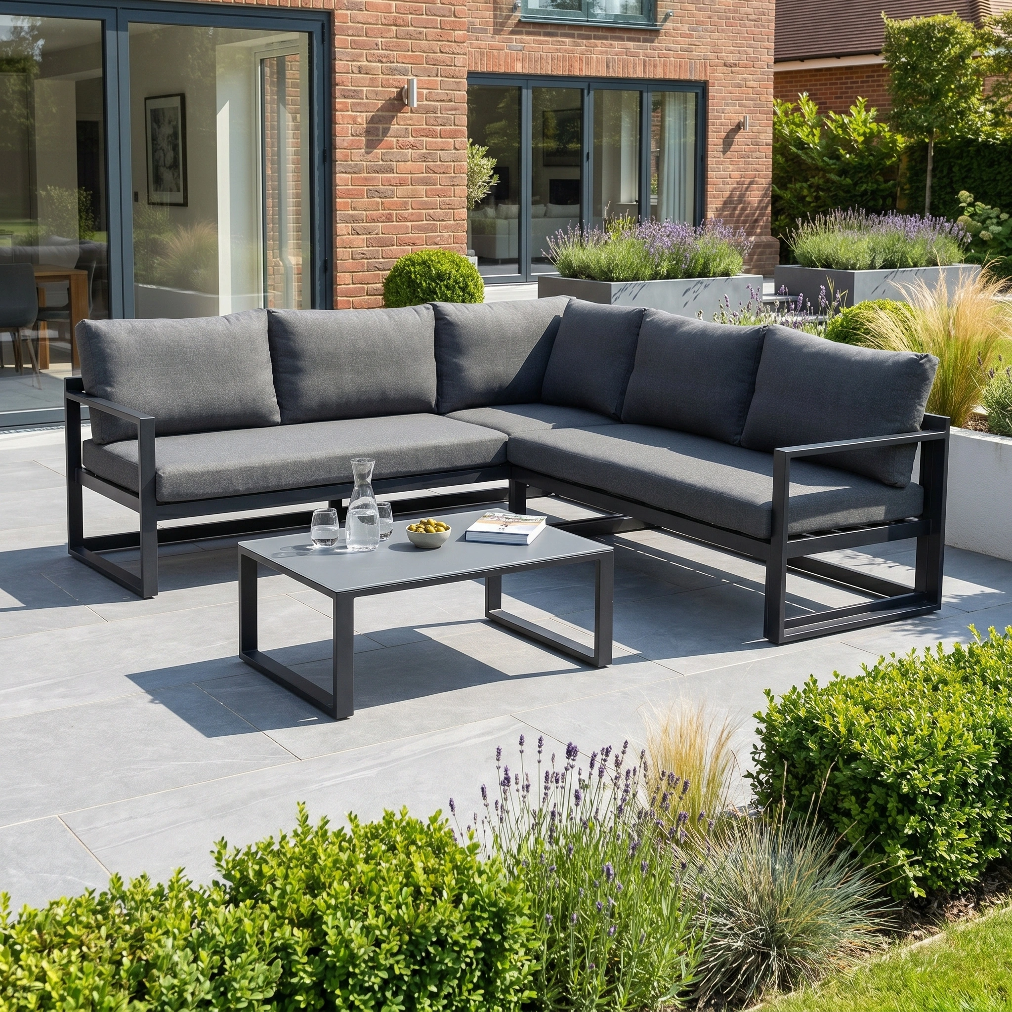 The Havana Corner Group Set with Reclining Feature and Coffee Table in Charcoal is ideal for relaxing in style on your patio, offering modern comfort as outdoor living furniture surrounded by a landscaped garden.