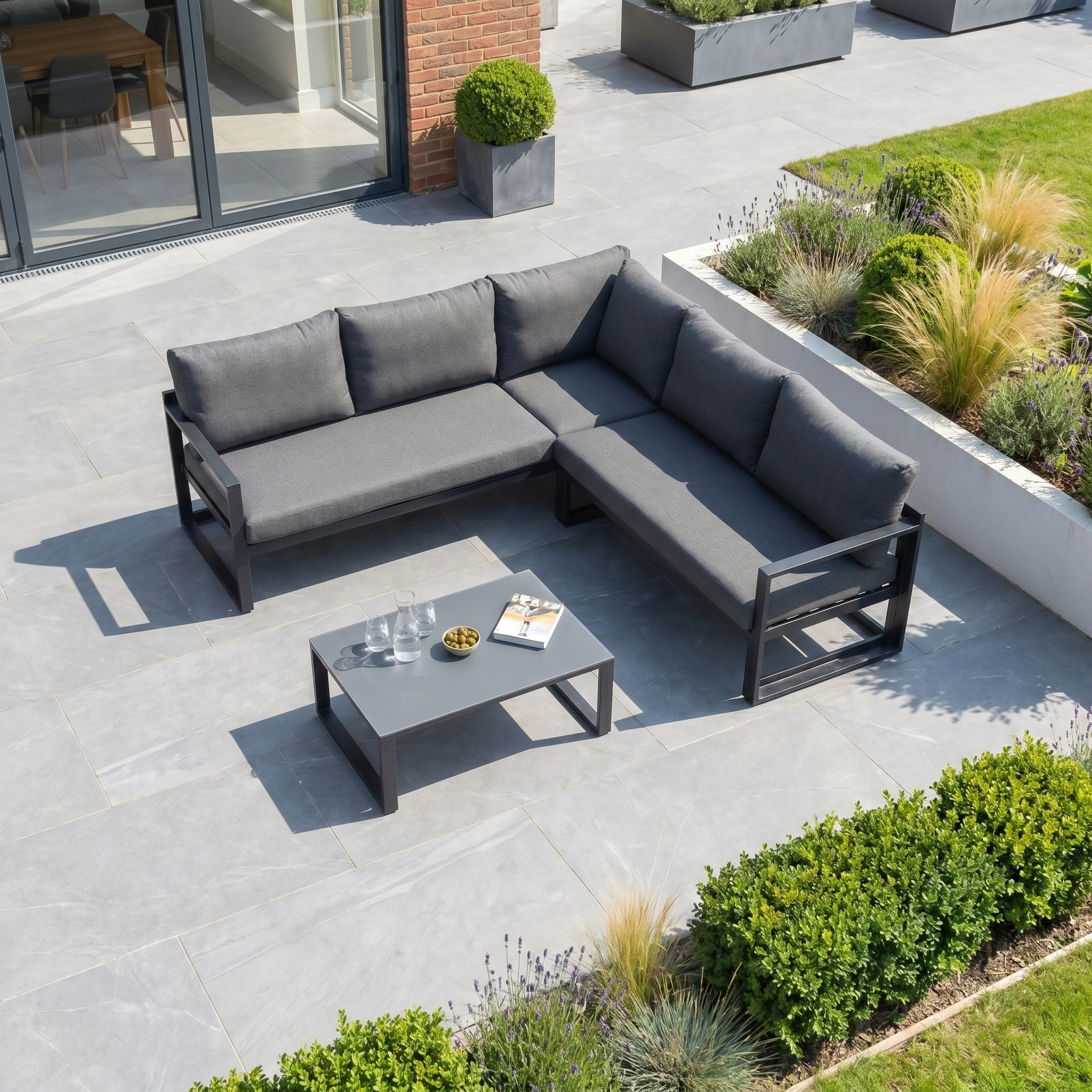 Modern patio featuring the Havana Corner Group Set with Reclining Feature and Coffee Table in Charcoal, complete with drinks and snacks, surrounded by lush greenery—ideal for relaxing with premium outdoor living furniture.