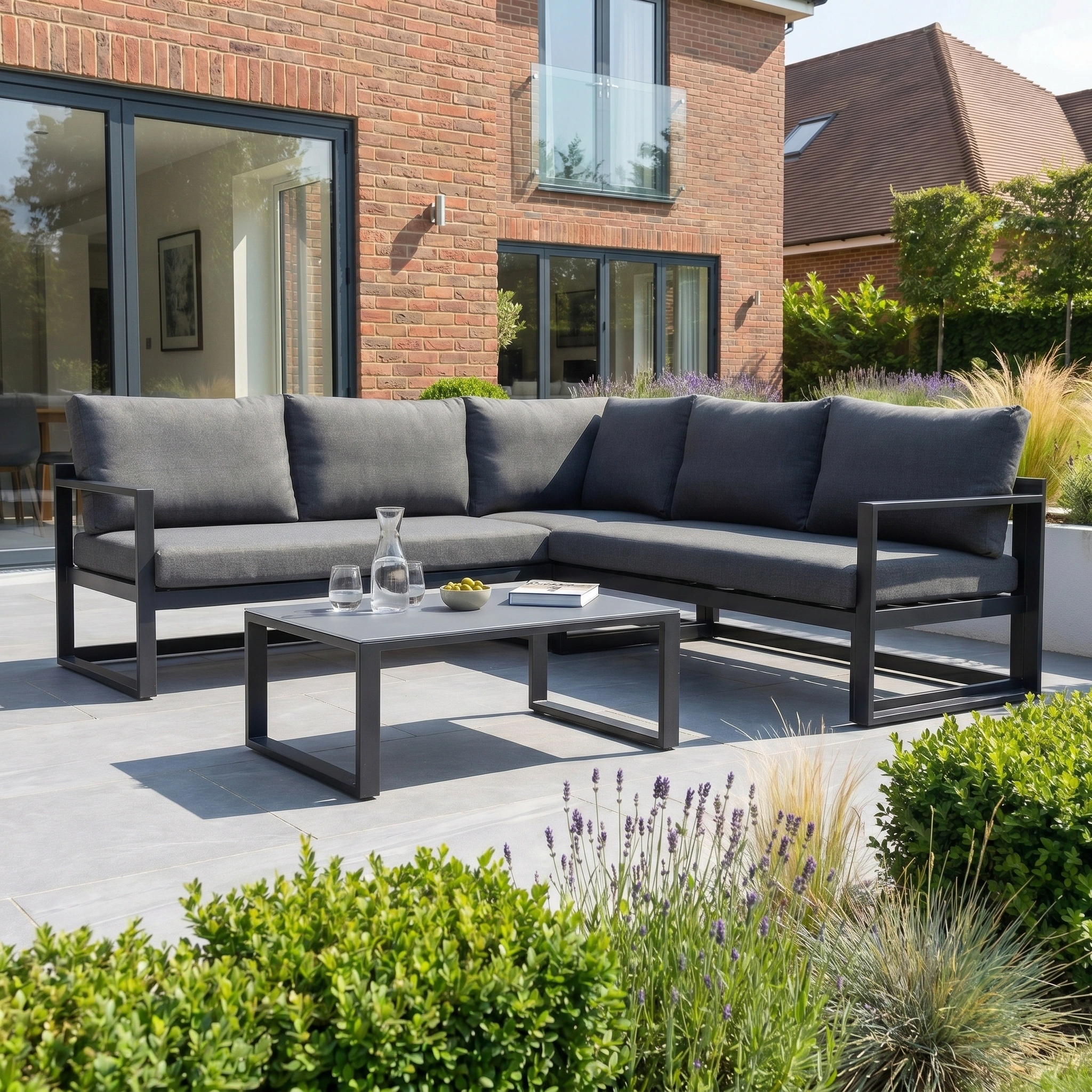 Create a modern outdoor patio with the Havana Corner Group Set with Reclining Feature and Coffee Table in Charcoal—stylish, versatile furniture perfect for relaxing on your terrace beside a brick house.