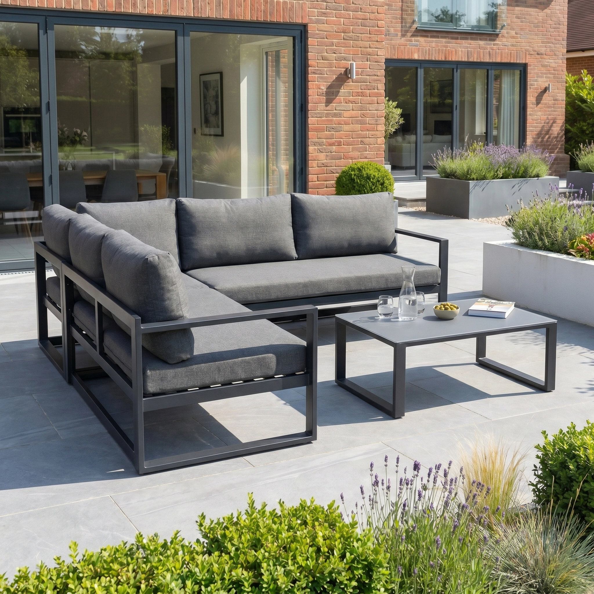 Modern outdoor patio with the Havana Corner Group Set in charcoal, featuring a reclining sectional sofa and coffee table, set before a garden and brick house.