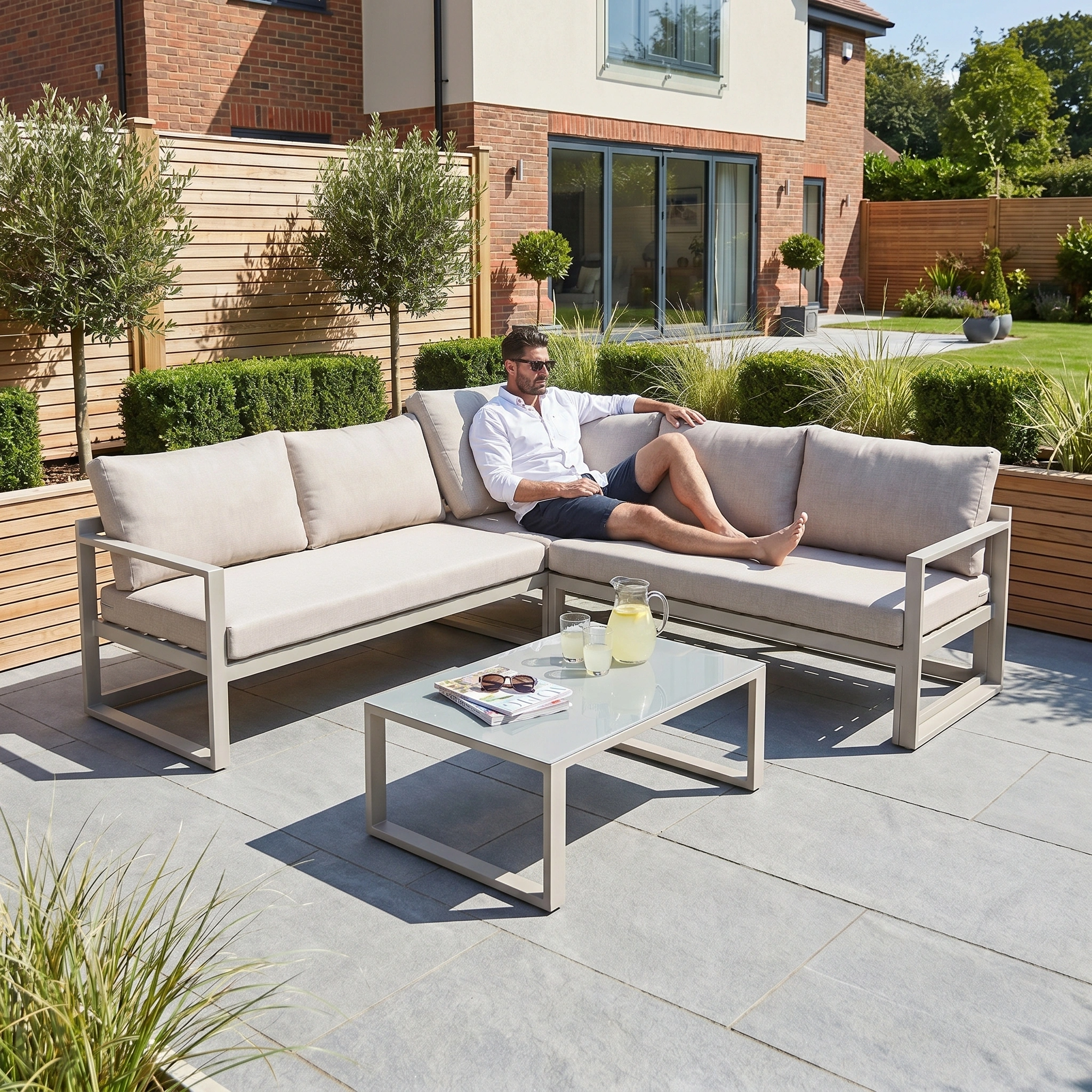 A man relaxes on the Havana Corner Group Set with Reclining Feature and Coffee Table in Latte, enjoying a sunny garden with lemonade and magazines on the table in front of him.