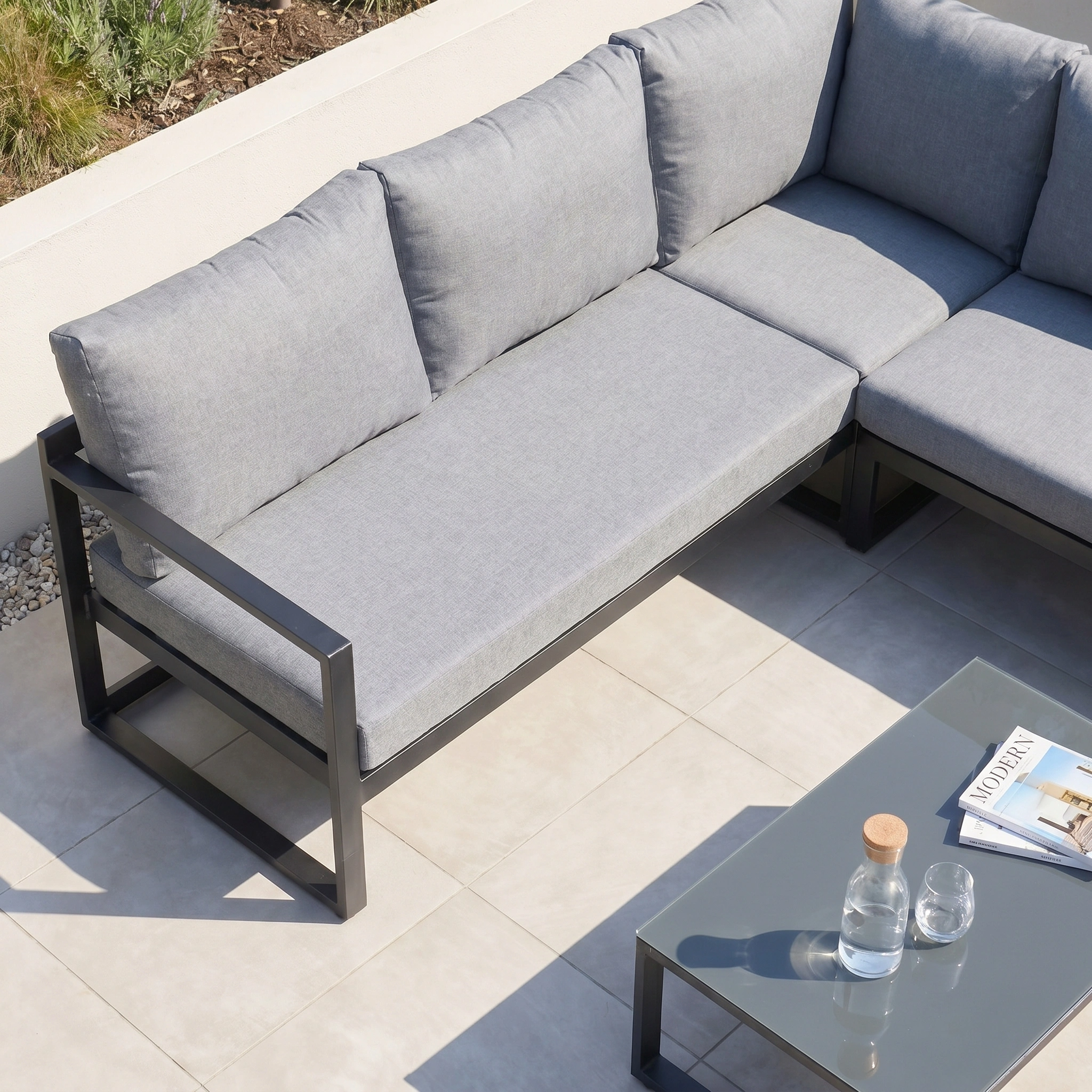 The Havana Corner Group Set with Reclining Feature and Coffee Table in Washed Grey offers comfort and style for outdoor relaxation, shown here on a tiled patio with a magazine and water carafe on the coffee table.