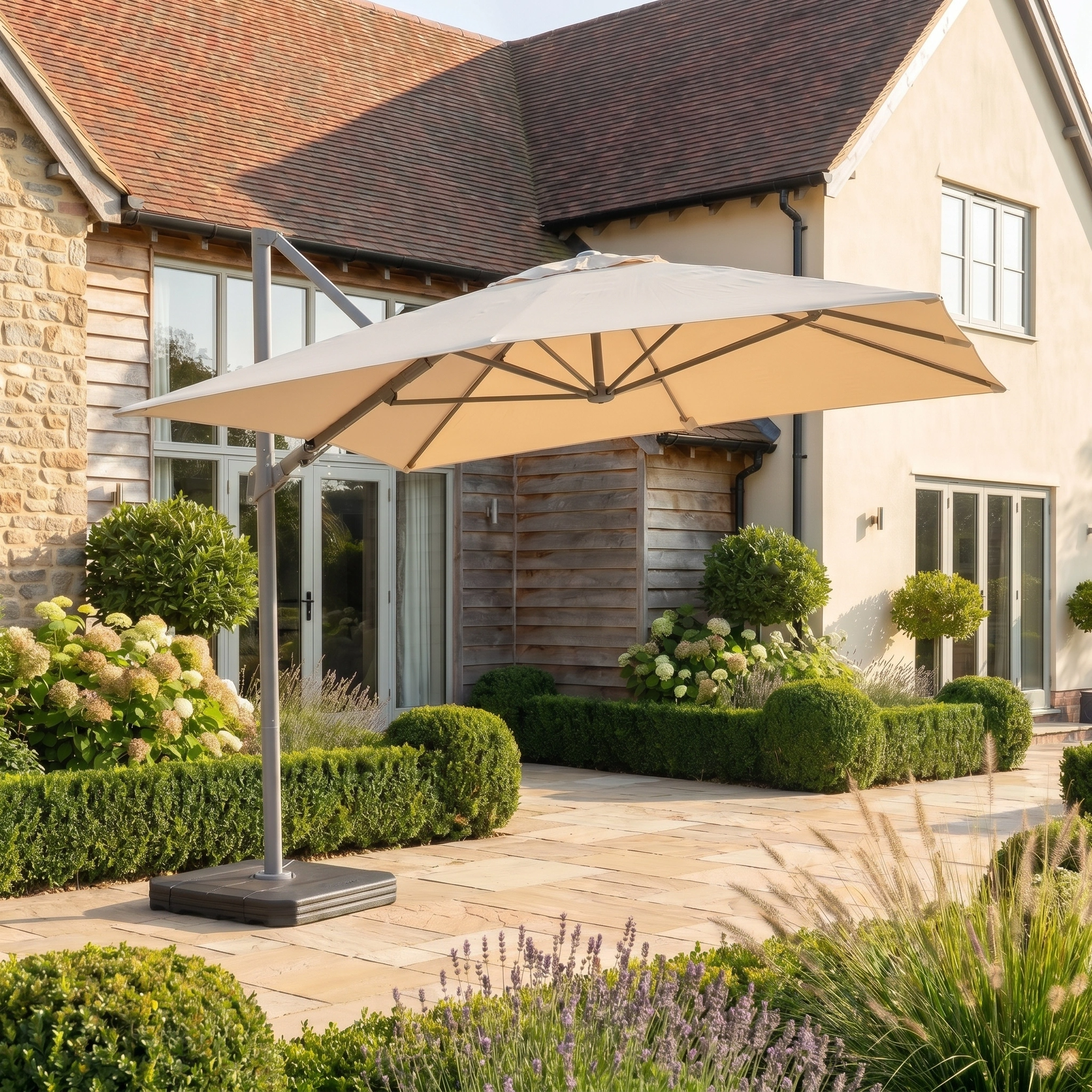 The Hera 2.7m Square Cantilever Parasol with LED Lighting in Beige shades a stone terrace by a modern house, features UV50+ sun protection, and complements lush green landscaping.