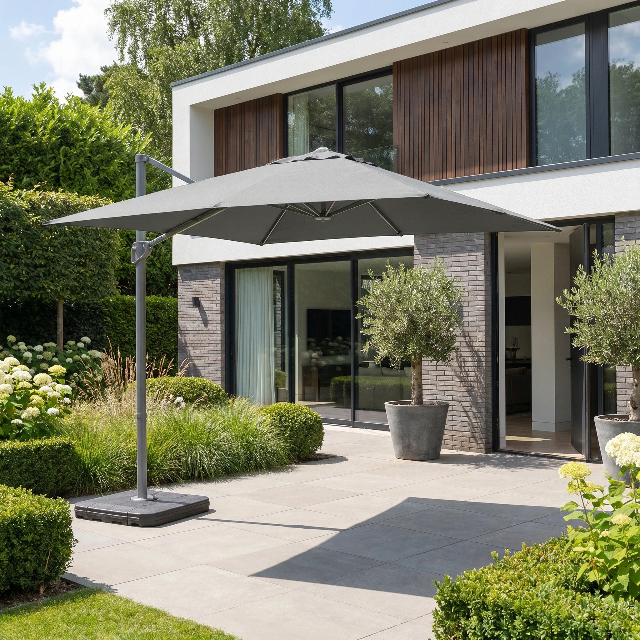 The Hera 2.7m Square Cantilever Parasol with LED Lighting in Charcoal shades a modern tiled patio, surrounded by potted plants and lush greenery.