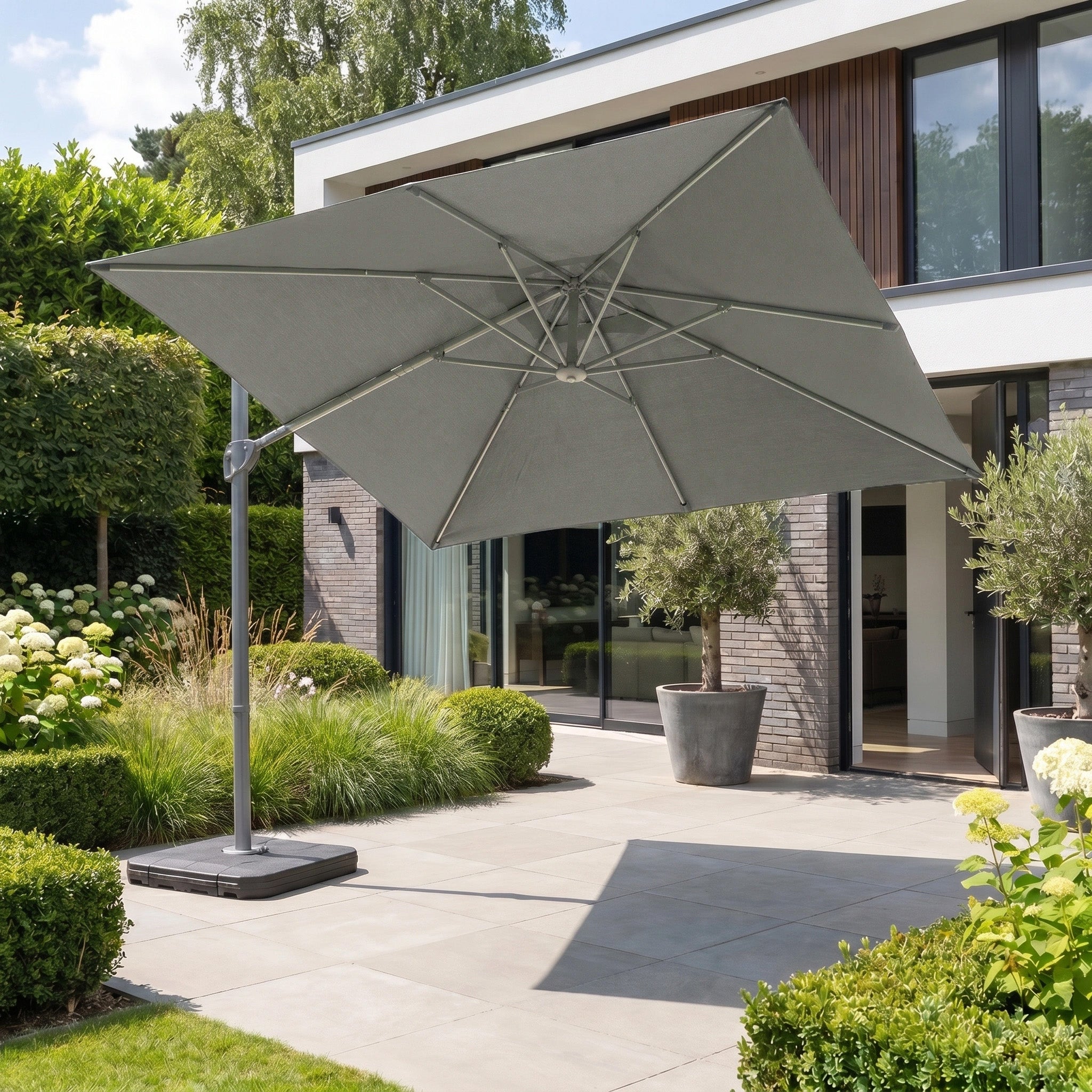 The Hera 2.7m Square Cantilever Parasol with LED Lighting in Charcoal shades a modern outdoor area with plants beside a contemporary house.
