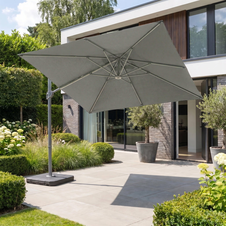 The Hera 2.7m Square Cantilever Parasol with LED Lighting in Grey stands open on a modern backyard patio near glass doors and lush plants.