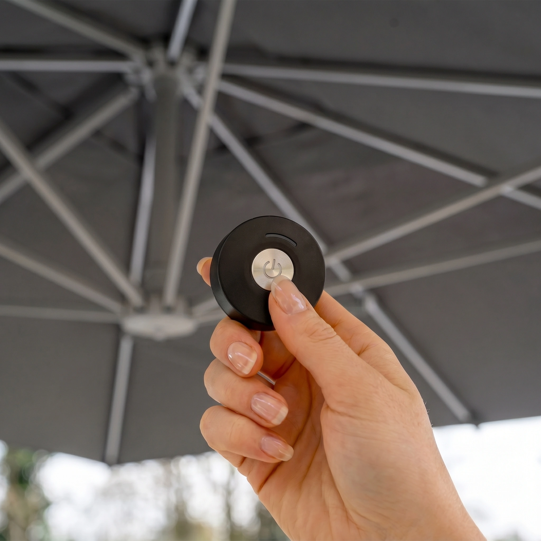 A hand holds a small black remote under the Hera 3m x 2m Rectangular Cantilever Parasol with LED Lighting in Charcoal, featuring a UV50+ spuncrylic canopy.