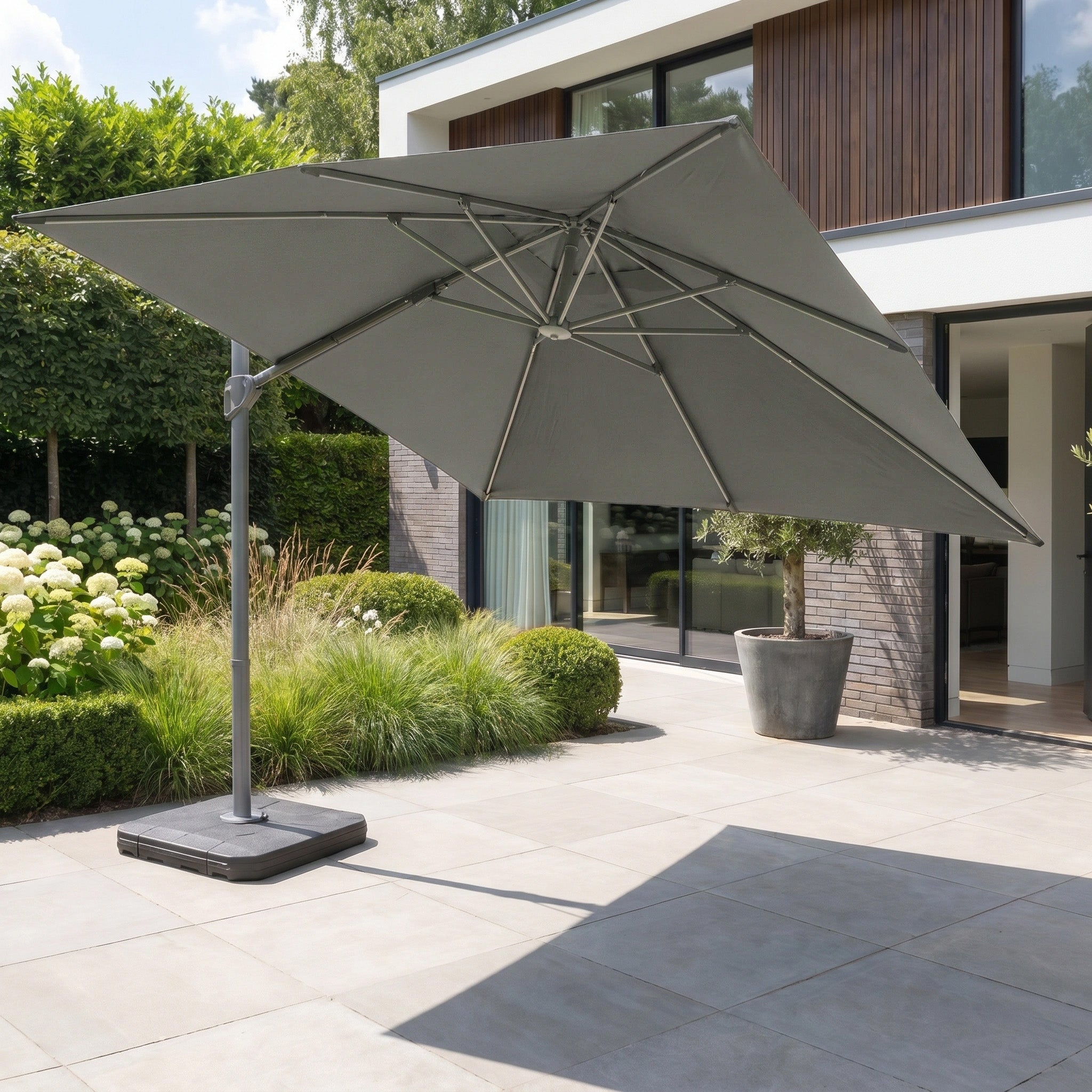 The Hera 2.7m Square Cantilever Parasol with LED Lighting in Charcoal is shown on a modern tiled terrace with plants, set against a contemporary house backdrop.